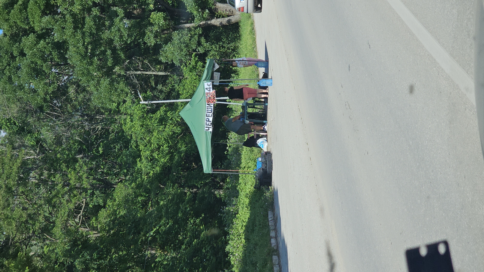 The image depicts an outdoor roadside stall selling cherries. A green canopy tent is set up, under which a table and several people are visible. A man in a dark shirt and shorts is standing behind the table, appearing to be engaged in work. A woman in a black top and pink shorts is also standing near the table. A young girl in a white and pink outfit stands to the right of the stall, holding a large blue water jug. The stall has a sign that reads "ЧЕРЕШИ 4€" (Cherries 4€), with an image of cherries. Various items are placed around the stall, including black crates, a white bag, and more blue water jugs. The stall is situated next to a paved road with a white line marking. Behind the stall is a dense line of green trees, suggesting a natural or semi-rural environment. A silver car is visible in the background to the right. The overall scene suggests a vendor operating along a road, likely in or near Karlovo, Bulgaria, given the Bulgarian text on the sign and the likely context of roadside fruit sales in the region.
