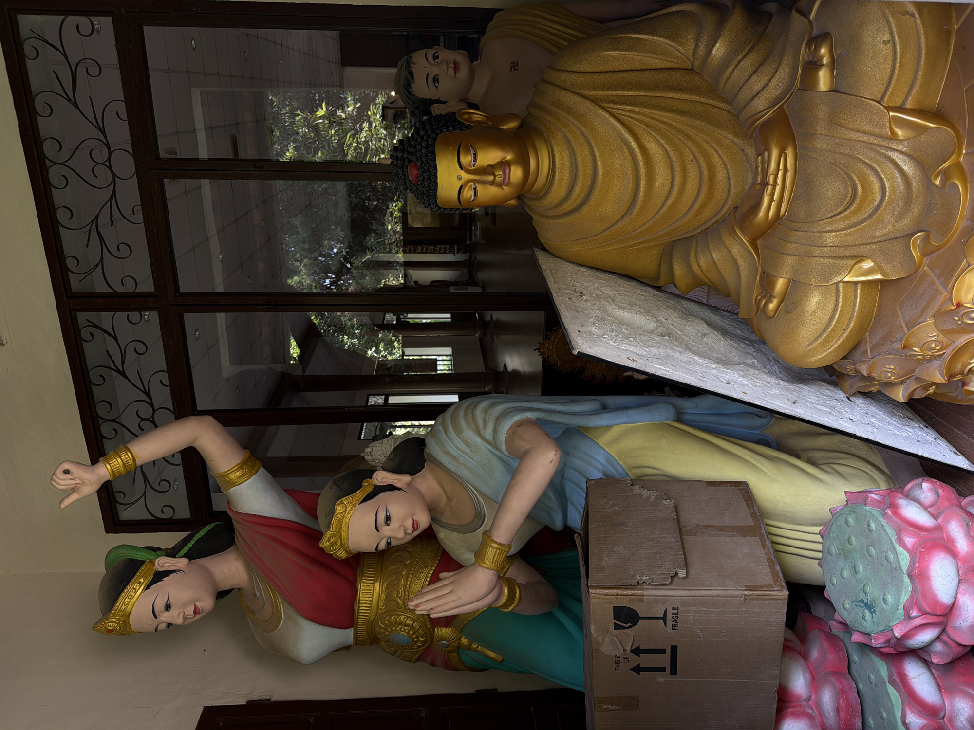The photograph, taken in Thủy Xuân Ward, Vietnam, shows several Buddhist statues. The largest is a golden Buddha in a seated meditative pose, placed near a window with decorative ironwork. Behind the Buddha is a smaller figure with a swastika symbol on its chest. In the foreground, two statues depicting female figures are positioned side-by-side. One figure extends her arm upwards, while the other clasps her hands together in a gesture of prayer. A cardboard box labeled "FRAGILE" partially obscures the second female figure and a decorative lotus base.