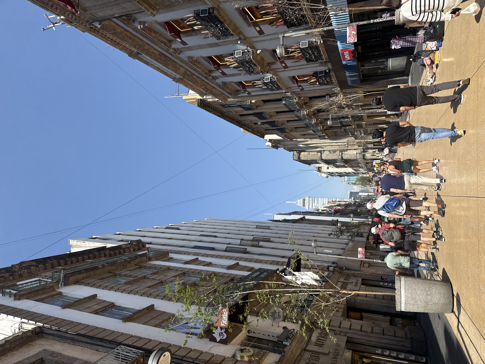 A street scene in Mexico City shows people walking along a pedestrianized street between buildings. The street is paved, and the buildings are of various architectural styles. The sky is blue and clear. The people are walking in the same direction. The environment appears to be urban and typical of a city center. The Torre Latinoamericana building is visible in the distance. A Levi's store is visible on the right.