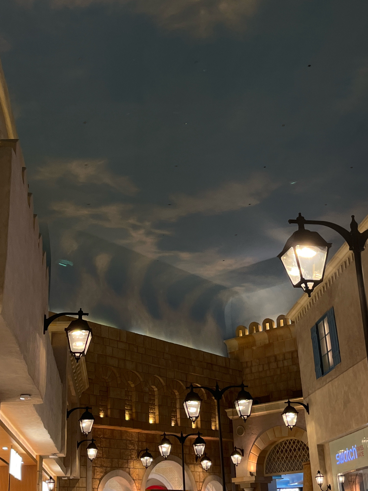 This is an indoor scene, likely in a shopping mall or entertainment venue in Dubai, United Arab Emirates, designed to resemble an old European street. The ceiling is painted with a cloudy sky motif, creating an illusion of being outdoors. Several lampposts with antique-style fixtures line the walkway, casting a warm light on the faux stone and stucco building facades. The architecture features arched doorways and windows, and some buildings have decorative crenellations. A sign for "Swatch" is visible on the right side, indicating a commercial establishment. There are no people visible in the frame. The overall atmosphere is one of artificial ambiance designed to evoke a specific historical or geographical setting.