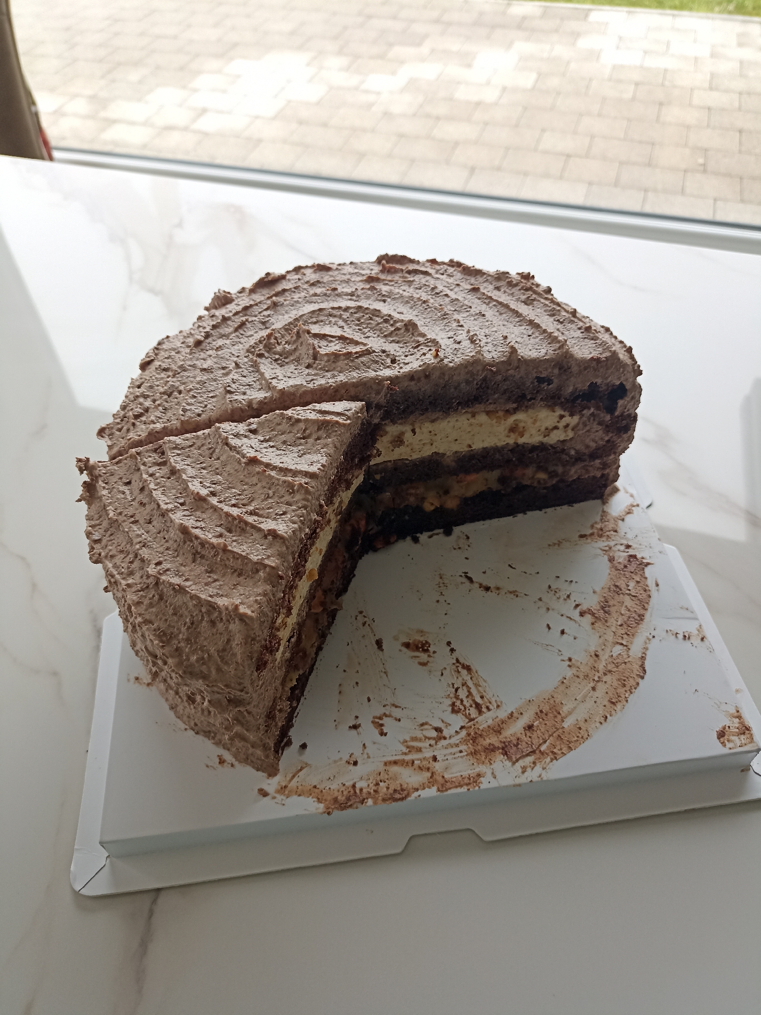 A chocolate cake with a slice removed sits on a white cardboard base, which is resting on a marble-patterned countertop. The cake is covered in textured chocolate frosting, resembling tree bark. The cut reveals multiple layers of dark chocolate cake interspersed with lighter colored cream and nut-filled fillings. Crumbs and smears of chocolate frosting are visible on the cardboard base and the countertop. In the background, through a window, a paved outdoor area with some greenery is visible. The lighting suggests an indoor setting, likely a home or a cafe. The image does not contain any information or context related to the location in Doirí Beaga, Ireland.