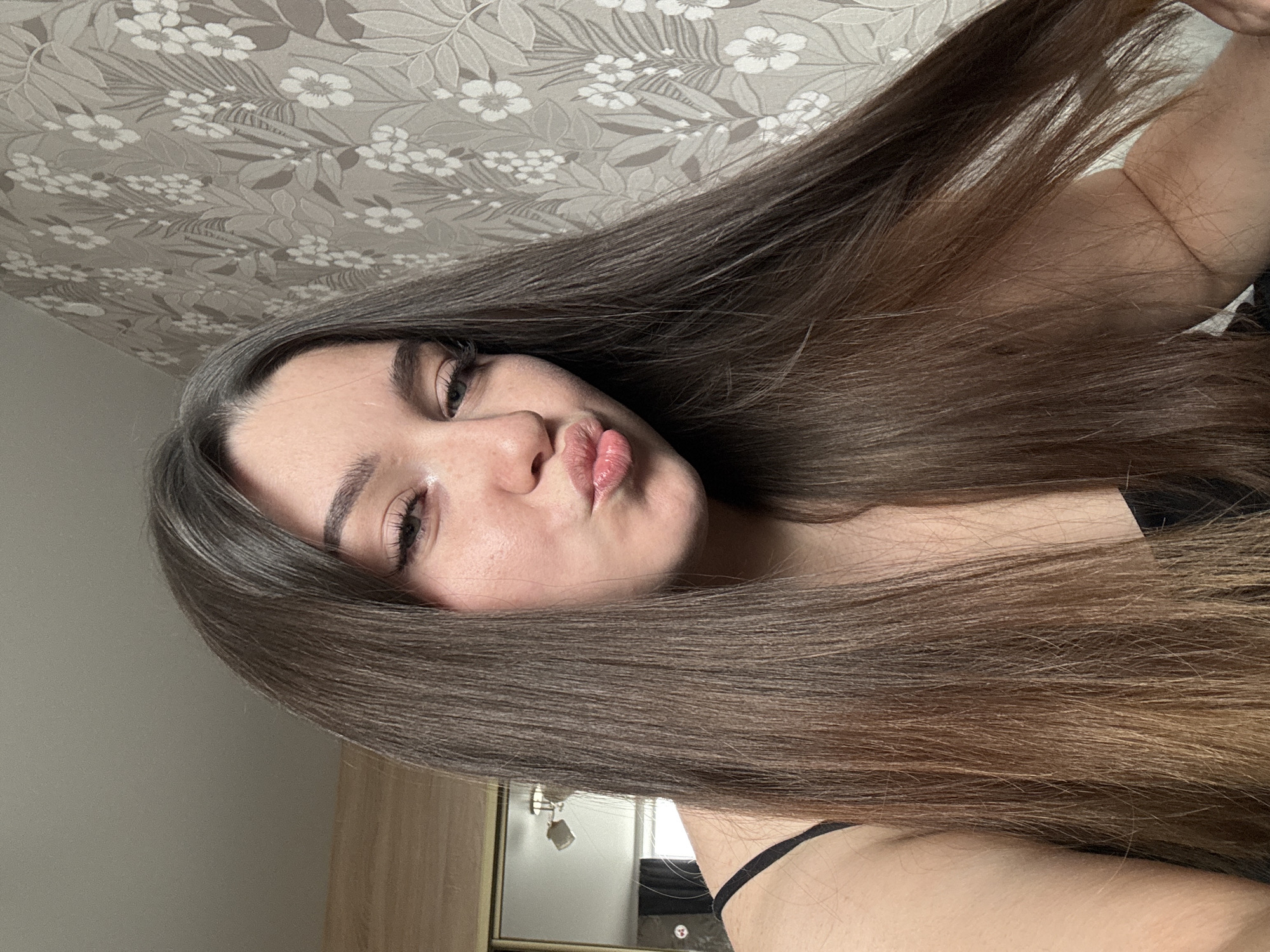 A woman with long brown hair and fair skin is posing for a selfie in an indoor setting. She is pouting her lips towards the camera. The background includes a floral wallpaper and a glimpse of what appears to be a kitchen area with cabinets. There is no indication that this image was taken in the city of Perm, Russia.