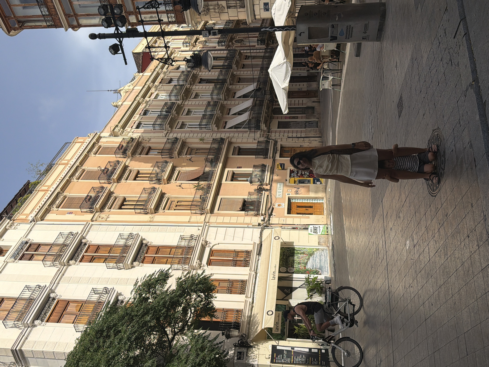 This media file captures a street scene in what appears to be a European city, possibly Spain, given the text "CALLE" and "CAU DE SERRANS". The image is oriented vertically, giving a sense of looking up at the buildings.
The primary subjects are people engaged in everyday activities. In the foreground, a woman is seen holding a small child, with the child seated on her shoulders. Further down the street, a man is interacting with a bicycle, possibly preparing to ride it or park it. In the background, several people are seated at an outdoor cafe, enjoying the day.
The scene is set along a cobblestone street lined with multi-story buildings featuring ornate balconies and large windows. There are awnings extending from some of the buildings, suggesting businesses, one of which is identified by the text "lavin snack&bar". A sign indicating "RENT A BIKE" is also visible, pointing to a potential tourist activity.
The time of day appears to be daytime, with bright sunlight casting shadows, suggesting a sunny day. The overall mood is one of a lively urban environment, with people going about their day in a pleasant atmosphere. The presence of trees adds a touch of nature to the built environment. The visible text provides specific location cues and identifies businesses operating in the area.
