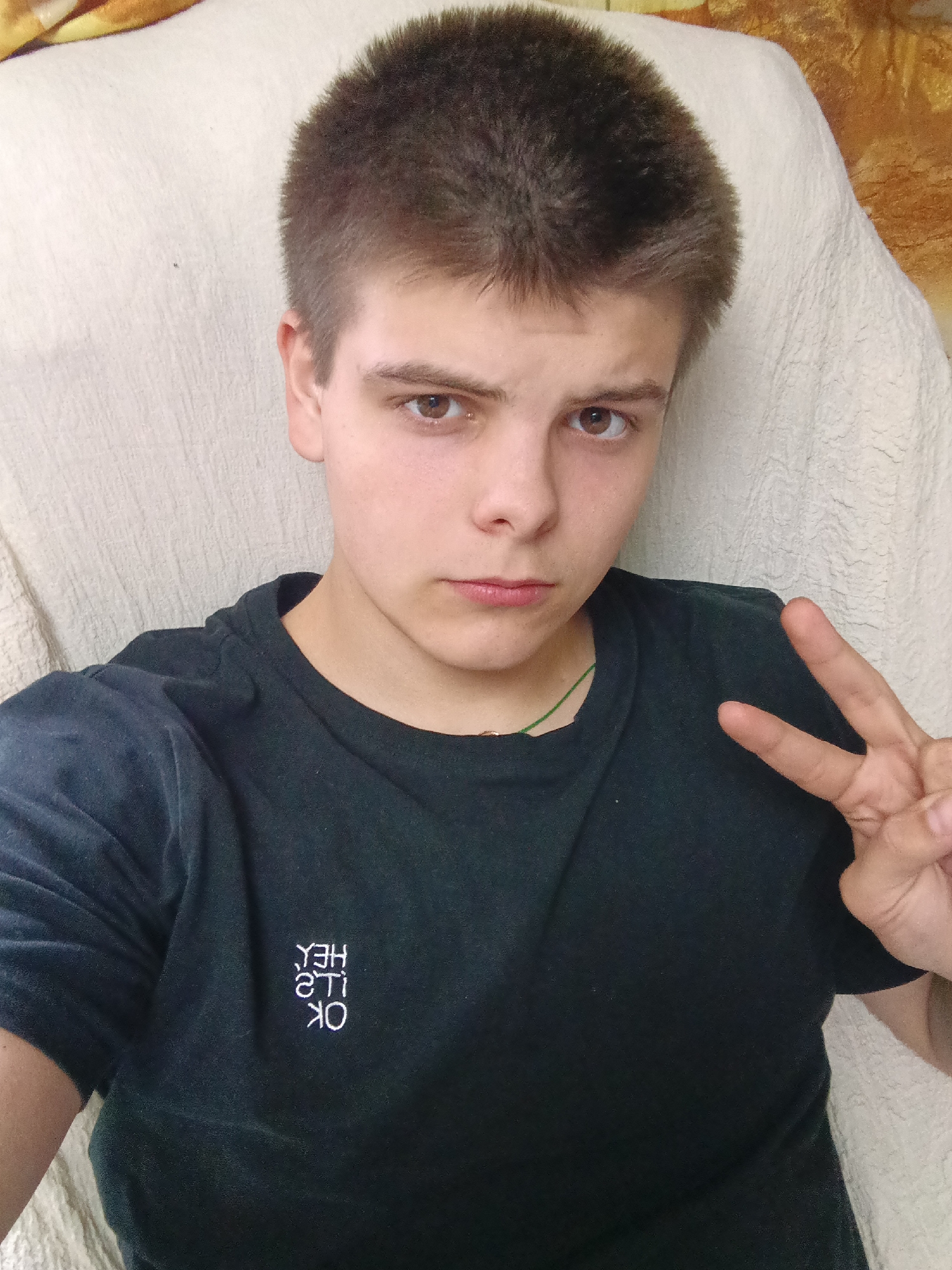 A fair-skinned person with short brown hair and brown eyes is centered in the image. They are wearing a dark t-shirt with the words "OK, it's Hey" vertically stacked on the left side of the shirt. The person is making a peace sign with their right hand. The background consists of a beige upholstered surface. Location: Unlikely Красный Маяк, Russia is in the image.
