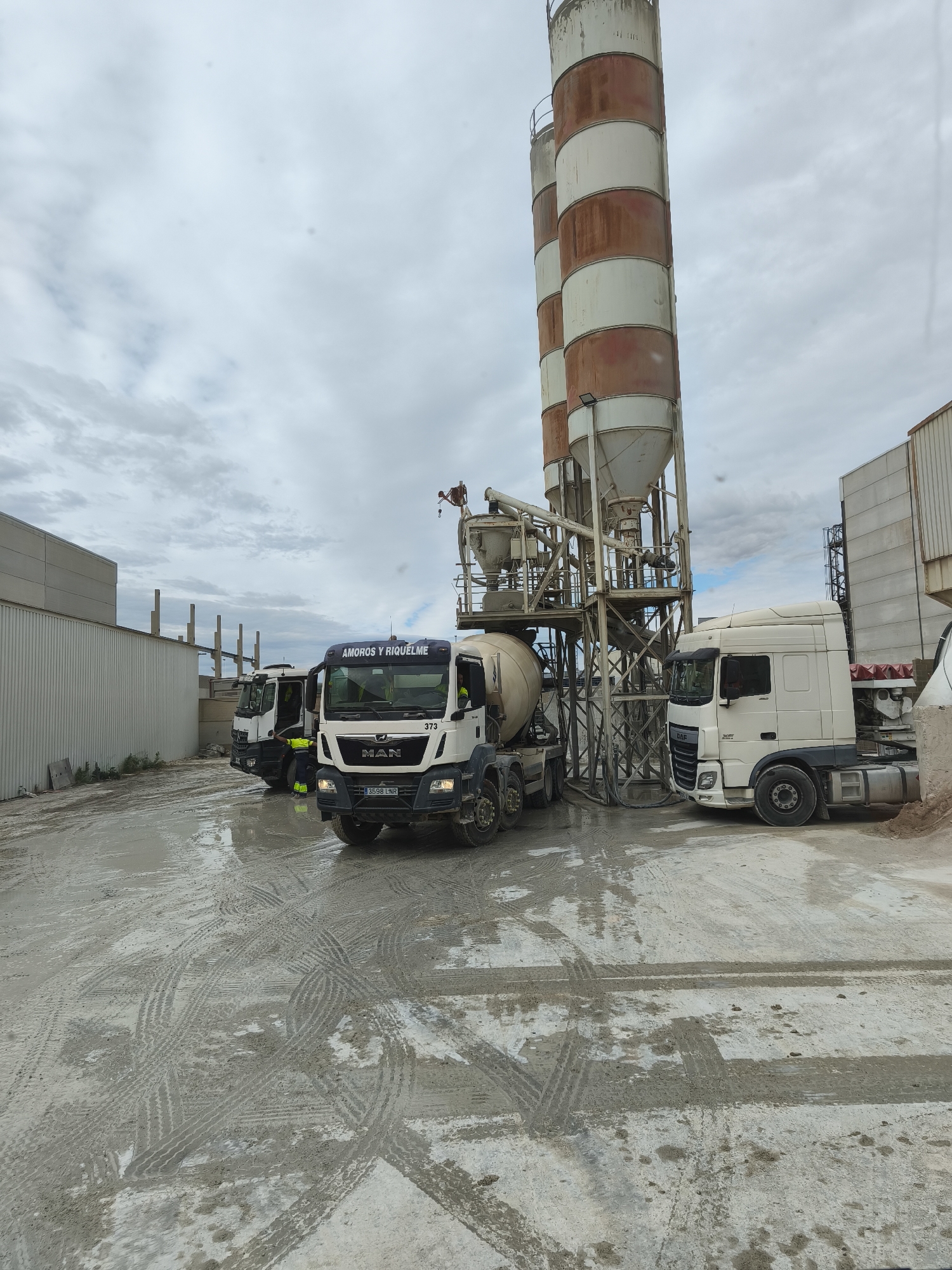 The image shows a construction site in Nàquera / Náquera, Spain. Several trucks are present, including a MAN cement mixer truck with license plate 3598 LNR, a DAF truck, and another truck partially visible. A tall concrete mixing tower stands behind the trucks. The ground is muddy, with tire tracks visible. The sky is overcast. A person is working on the mixing tower. Several industrial buildings are visible in the background.