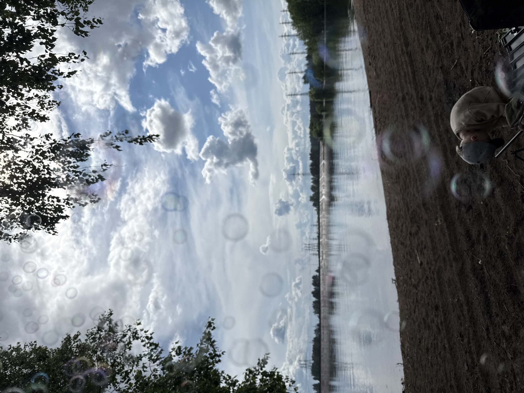 The image shows a landscape with a body of water, likely a river or lake, reflecting the cloudy sky. A dark patch of land is visible on the right side of the image, with sparse vegetation or perhaps dirt. A person, wearing a hat and a light-colored hoodie, is near the edge of the land. Bubbles are scattered throughout the image, giving the impression of someone blowing bubbles. Some trees are visible along the edge of the water and in the foreground. The presence of power lines in the background suggests an urban or semi-urban environment.
The location is Noviy, Russia.