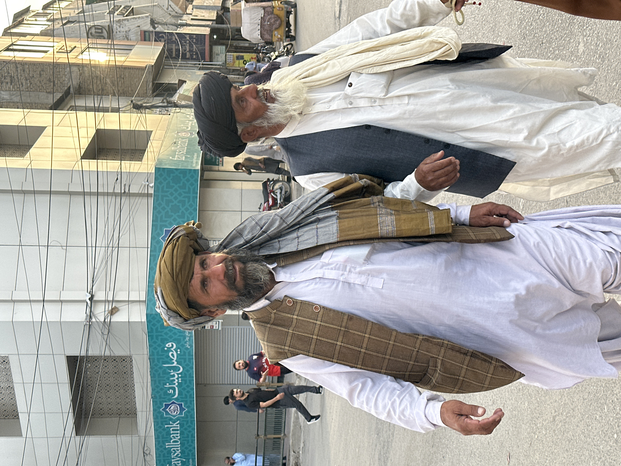 Two Pashtun men walk down a street in Quetta City Tehsil, Pakistan. The man on the left wears a brown plaid vest over a white shalwar kameez, a turban, and a shawl. The man on the right wears a dark vest over a white shalwar kameez, a black turban, and a cream-colored shawl. A bank sign with "Faysal Bank" written in English and Arabic is visible in the background, along with other buildings and street activity.