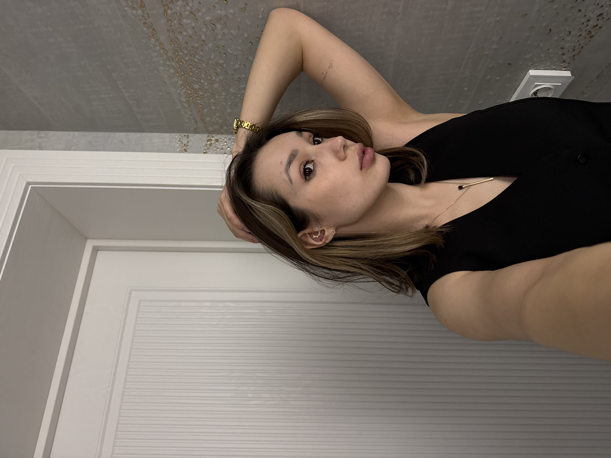 A woman with shoulder-length brown and blonde hair is lying down, looking at the camera. She is wearing a black top and a gold watch on her left wrist. A gold necklace is visible around her neck. She has multiple piercings in her left ear, adorned with jewelry. Her right arm is raised, with her hand behind her head. The background consists of a textured gray ceiling with gold speckles, a white electrical outlet, and a white door with horizontal panels. The scene appears to be indoors.