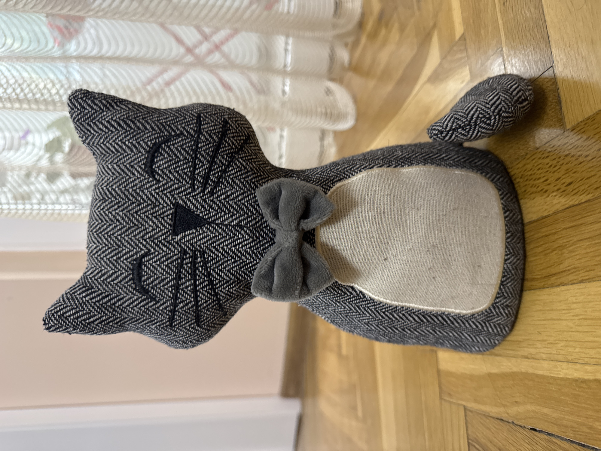 A decorative, stuffed cat toy lies on a wooden floor. The toy is gray with a herringbone pattern on the head and body, a gray bow tie, and a light gray patch on the body. The cat toy is near a white wall and a white curtain. The location is not discernible from the image, and I cannot ascertain if it is in Plovdiv, Bulgaria, based on the visual information.