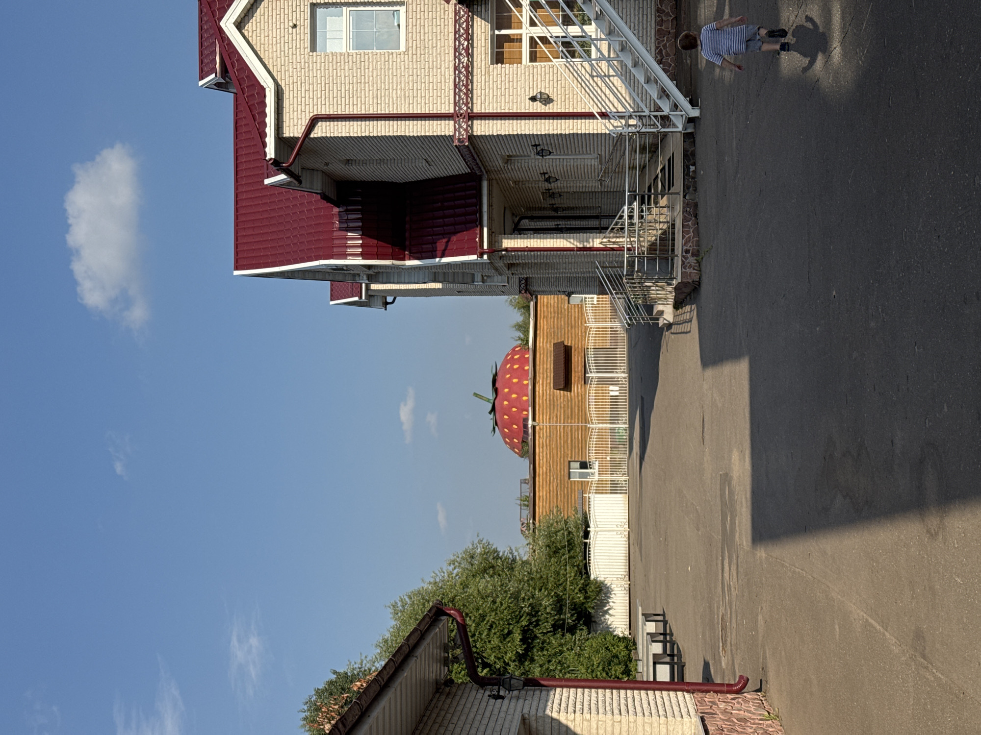 In the city of Sovkhoza imeni Lenina, Russia, a young boy with brown hair, wearing a blue and white striped t-shirt and grey shorts, walks away from the camera on a paved area. To the right of the boy is a light-colored brick building with a red metal roof and a white staircase leading to an entrance. The building has multiple windows. In the background, there is a wooden building topped with a large, red, strawberry-shaped dome with yellow seeds and a green stem. A white fence with an arched design runs in front of the wooden building, and beyond that, lush green trees are visible. To the left of the image is another light-colored brick building with a red metal roof and a gutter system. The sky is clear blue with a few small white clouds. The ground is asphalt with some light-colored patches and shadows cast by the buildings.