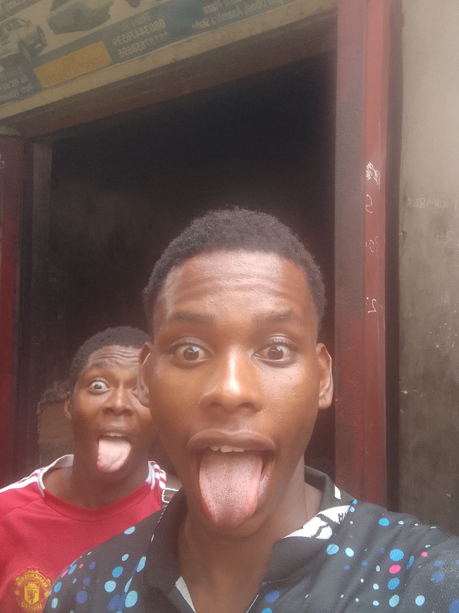 Two young men are sticking out their tongues and looking at the camera. One is wearing a red Manchester United jersey and the other a black shirt with blue and pink dots. They are in front of a building with a metal frame around a doorway. The building appears to be in Onitsha, Nigeria. There is an advert above the doorway.