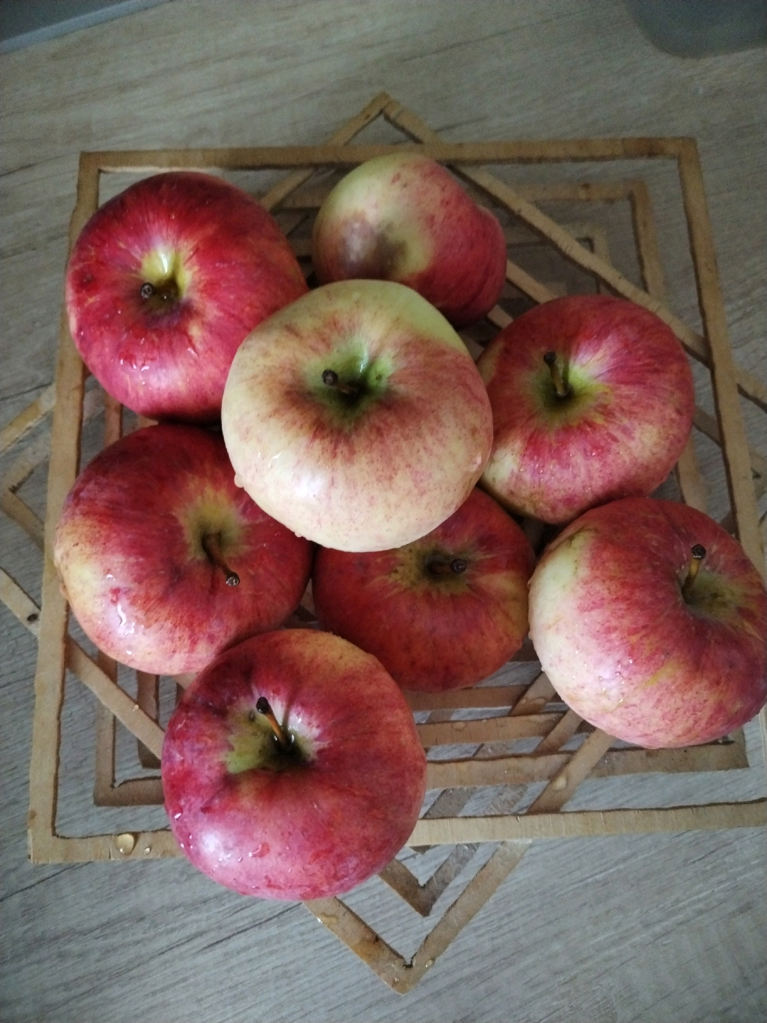 The image shows a close-up, overhead view of eight apples arranged in a woven wooden basket on a light brown wooden surface. The apples are primarily red with some yellow and green undertones, and appear to be freshly washed, with visible water droplets on their skins. The basket is square with an open weave design. The wooden surface has a grain pattern. There is no indication of the city of Baranavichy, Belarus in the image.