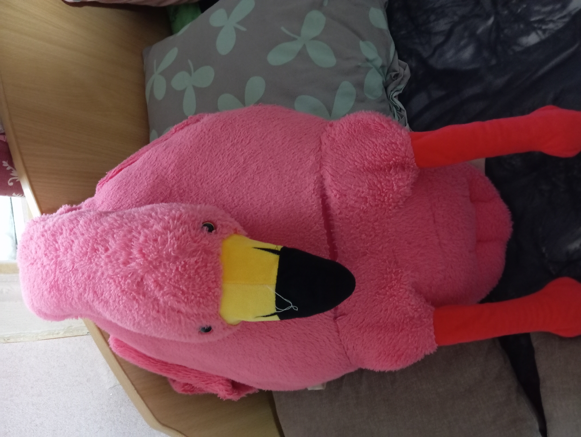 A plush pink flamingo toy is positioned upright on a bed. The toy features a yellow beak with a black tip, two small black eyes, and two red legs. It is situated next to a grey pillow with a clover pattern. The background includes a wooden headboard and a white textured wall. The scene appears to be indoors, likely a bedroom. The image does not contain information that places it in Barnaul, Russia, or any specific city.