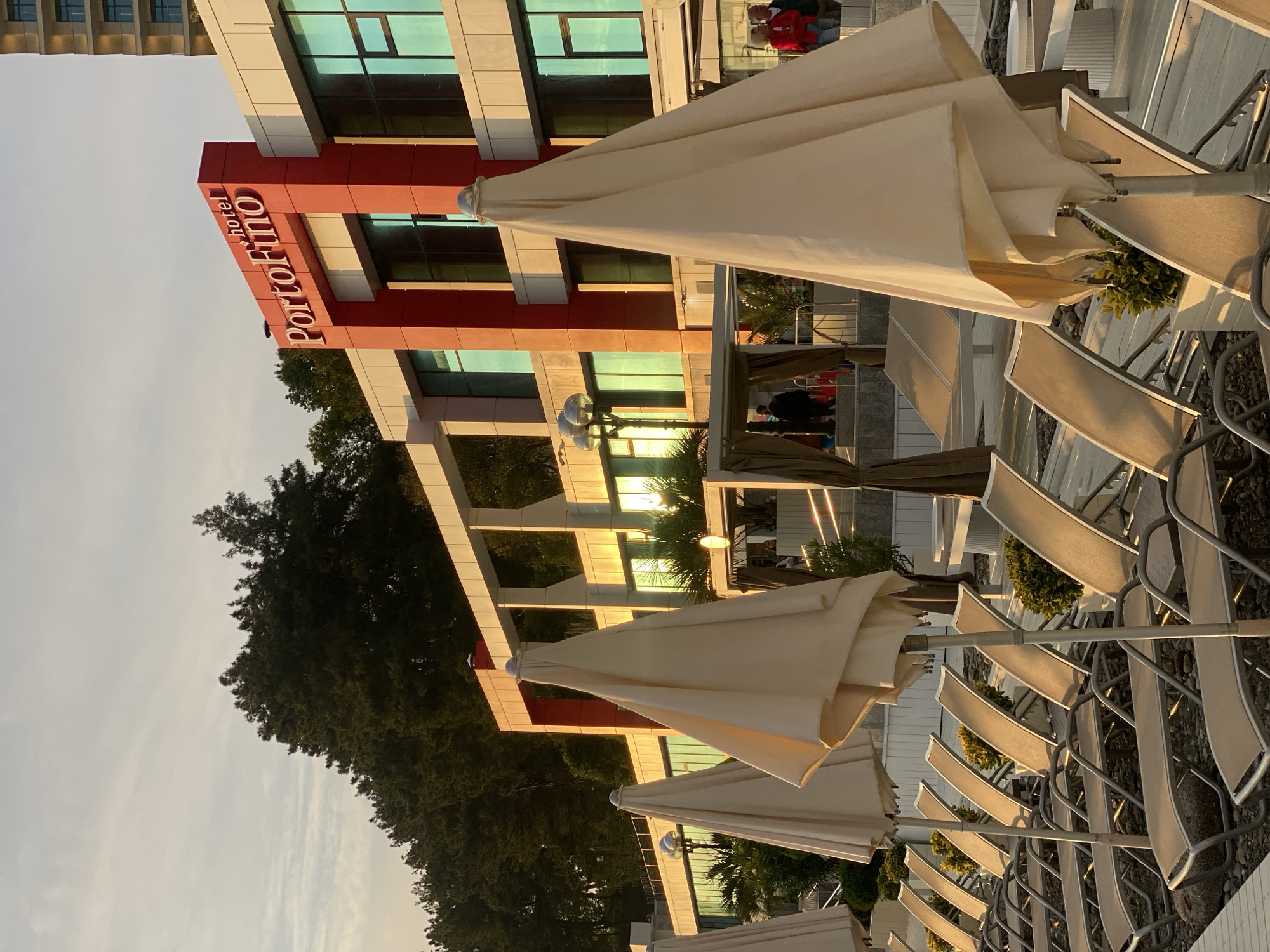 The image shows the exterior of the "Hotel Portofino" in Sochi, Russia. The building has a modern design with red and white accents and large windows reflecting the sky. Multiple folded, beige umbrellas are in the foreground, lined up near lounge chairs, likely on a patio or deck area of the hotel. Trees are visible behind the hotel. The sky appears to be a light, hazy color, indicating either morning or late afternoon lighting.