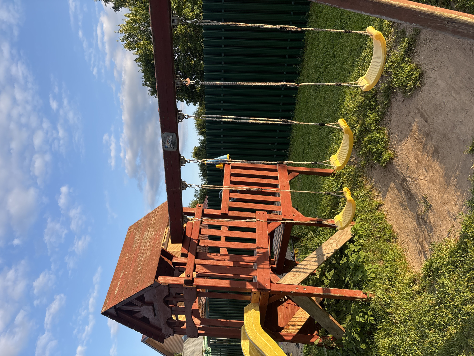 In Мещёры, Russia, a wooden children's playground set stands outdoors on a grassy area. The structure is red and features a red-tiled roof, a yellow slide, and a wooden ladder. Three yellow swings hang from a horizontal wooden beam. A green metal fence runs along the background. The sky is blue with scattered white clouds, and the sun is casting long shadows.