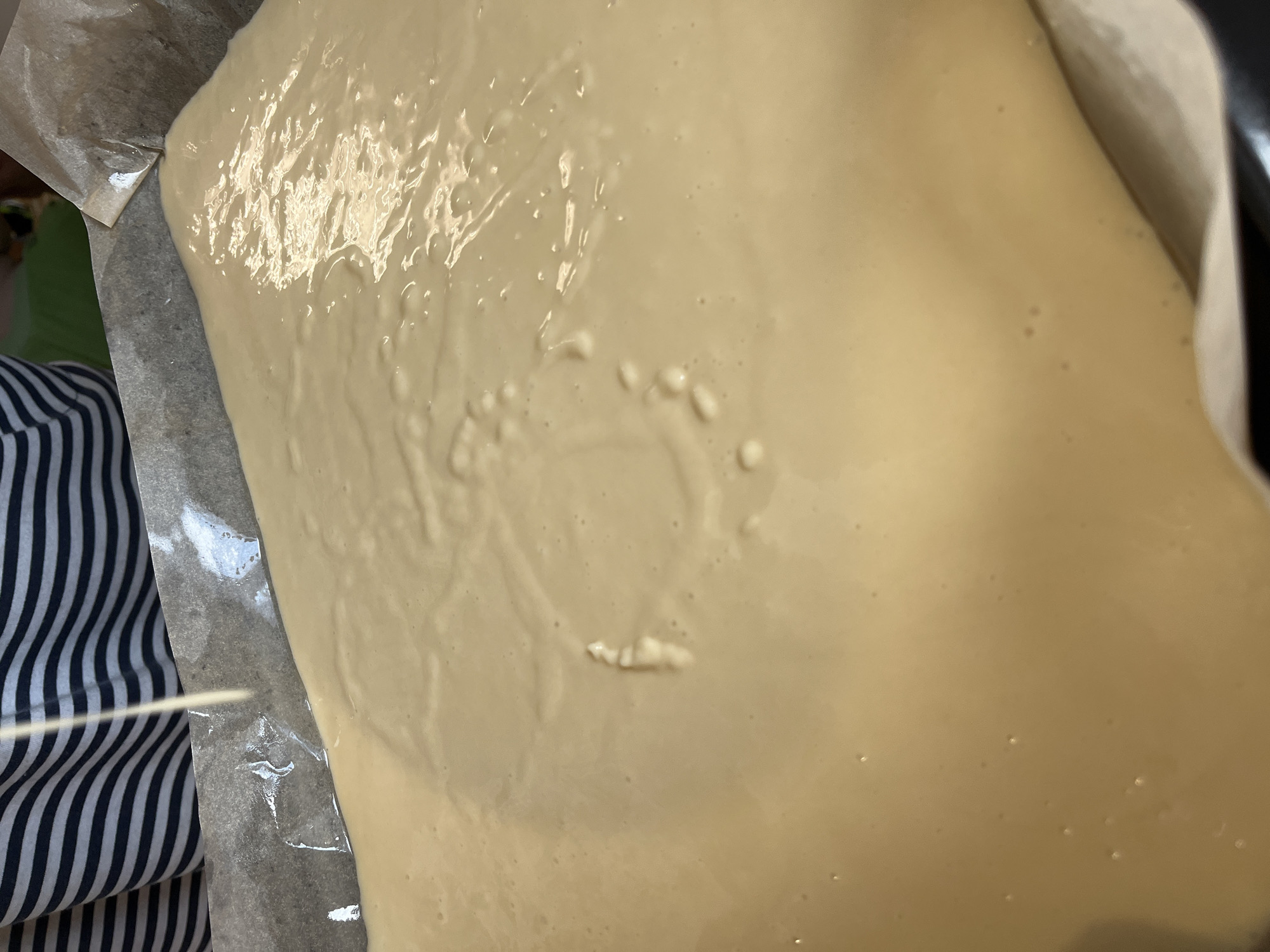 The image shows a close-up view of a light-colored batter being poured into a baking pan lined with parchment paper. A person wearing a striped shirt is visible in the upper left corner, with a thin stream of batter being poured from above into the pan. The batter is smooth and slightly glossy, with some small bubbles visible on the surface. The baking pan appears to be rectangular and is filled with batter up to about half its depth. The background is out of focus and appears to be a kitchen environment. There is no discernible information in the image that indicates a specific location in the city of Chaikovsky, Russia.