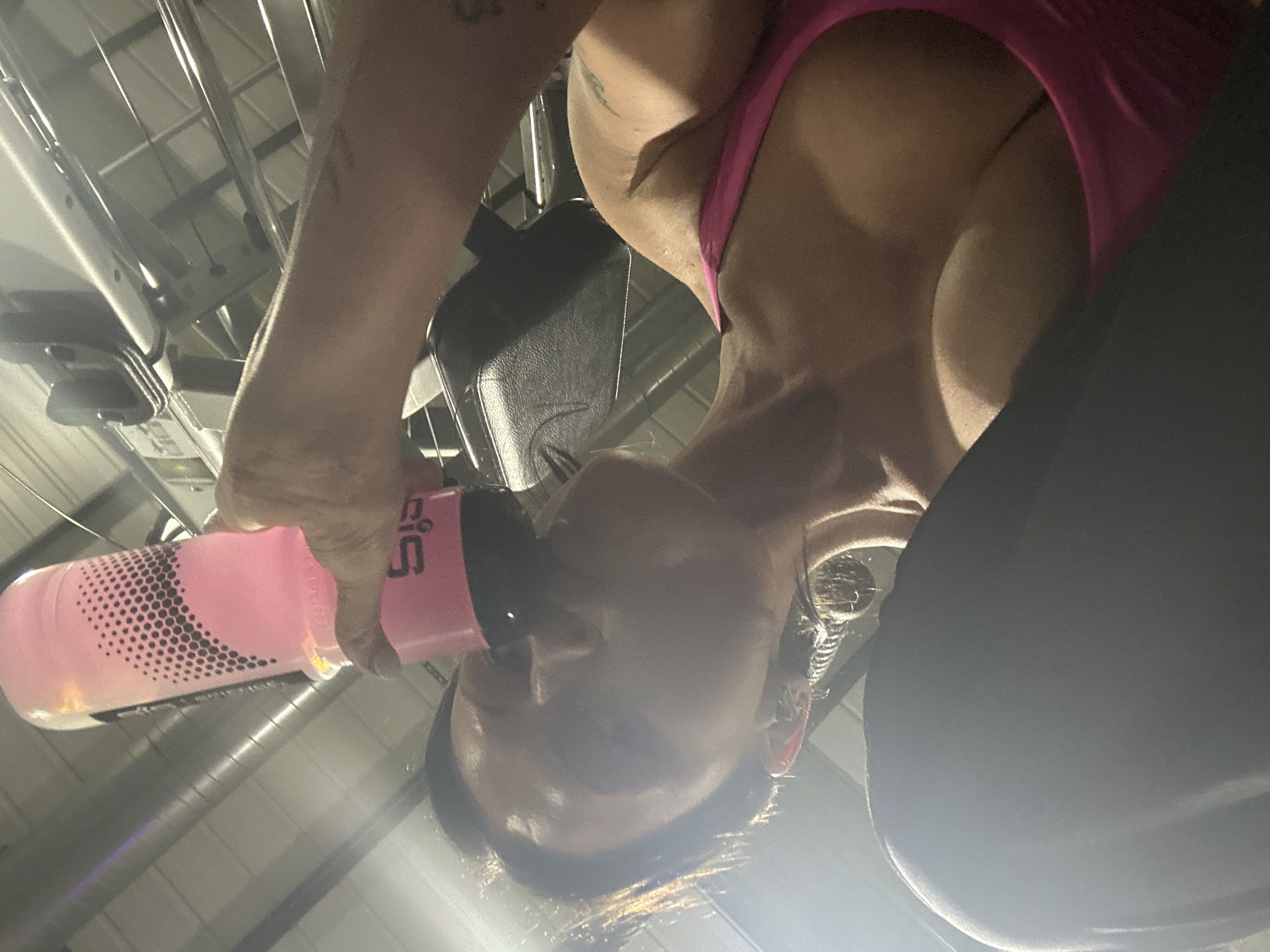 This image depicts an adult female mid-action, drinking from a pink Science in Sport (SiS) branded water bottle, held in her right hand. Her head is tilted upwards, and she wears a fuchsia athletic top and dark lower garments. A visible sheen indicates moisture on her skin. The background reveals industrial overhead structures, including parallel silver metallic bars and dark utility fixtures, alongside a partially visible black padded section of gym equipment positioned behind her right shoulder. The environment is an indoor fitness facility located in Tunbridge Wells, United Kingdom.