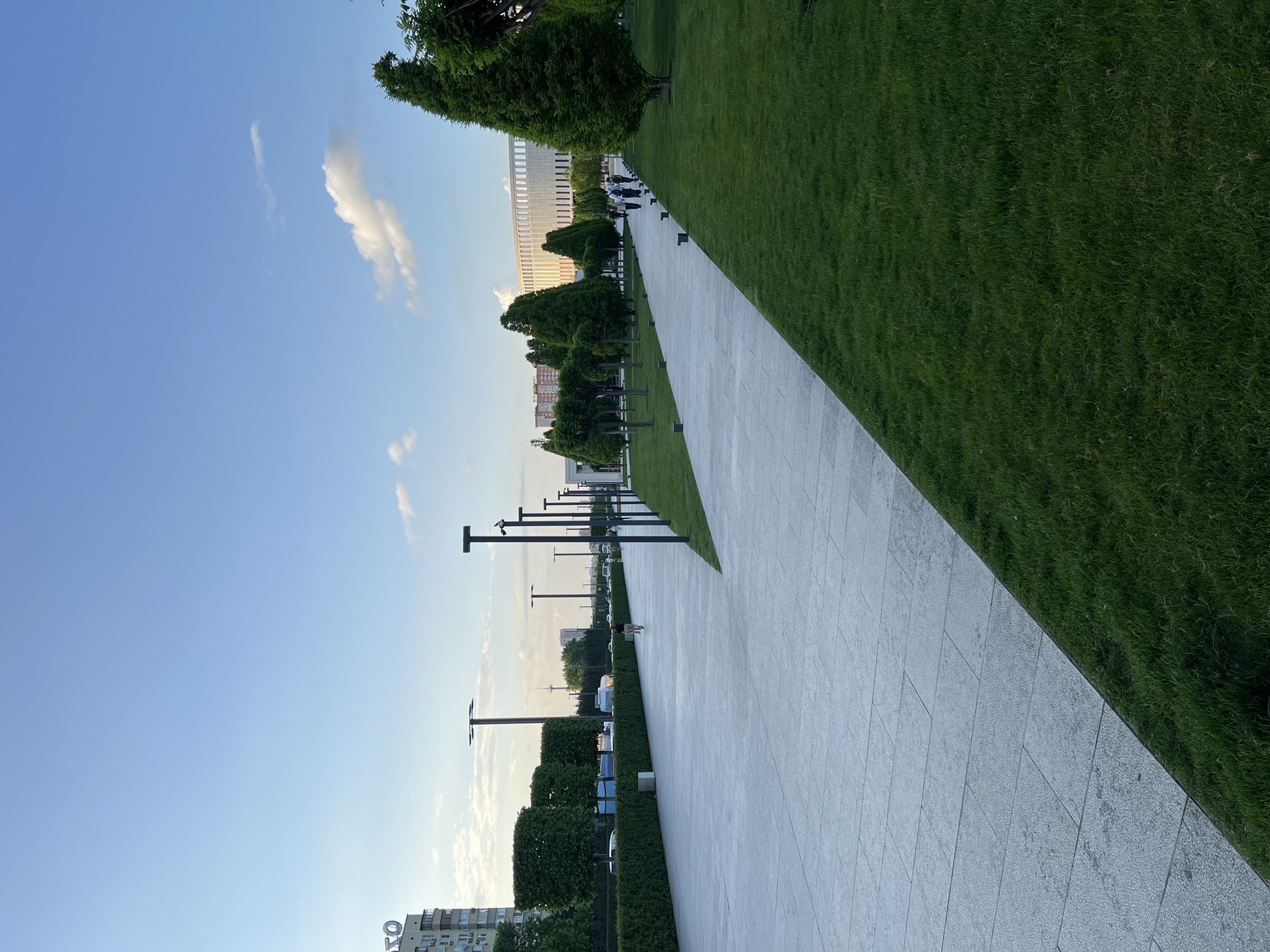 The image shows a paved walkway flanked by manicured green grass and trees in a park-like setting. A row of lampposts runs along one side of the walkway. In the background, there are buildings, including a prominent, large, light-colored structure that appears to be a stadium or large public building. The sky is blue with some scattered clouds. The image suggests the scene is likely in a modern or recently developed area. The presence of the FC Krasnodar Stadium (the large, light-colored building in the background) indicates the location is Krasnodar, Russia. No people are clearly visible in the foreground, but small figures that might be people can be seen in the distance along the walkway.