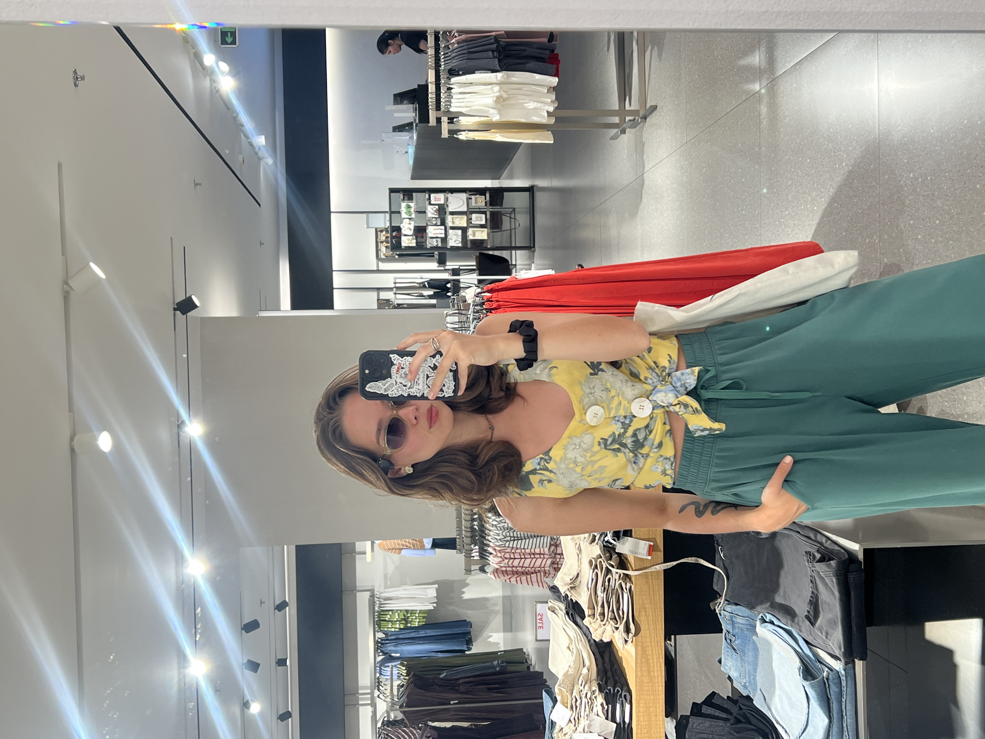A person wearing sunglasses and holding a phone with a patterned case is taking a mirror selfie in a clothing store. The person is wearing a floral yellow top and green pants. The background shows racks of clothing in a brightly lit retail environment. The reflection reveals a person in the distance browsing clothes. A rainbow reflection is visible in the upper left corner of the mirror. Given the presence of the "Zara" tag visible on clothing racks, this is a likely a Zara store. I do not have specific information to confirm that this is Nha Trang, Vietnam.