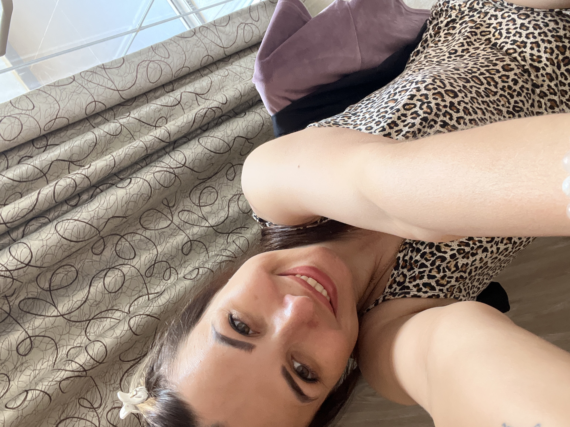 A woman with dark hair, a white clip in her hair, and a leopard print top smiles at the camera. She is leaning against a wall with patterned curtains behind her. Her left arm is raised, and her right arm is resting on her leg. A purple cloth is draped over her shoulder. The lighting suggests it is daytime, possibly near a window.
