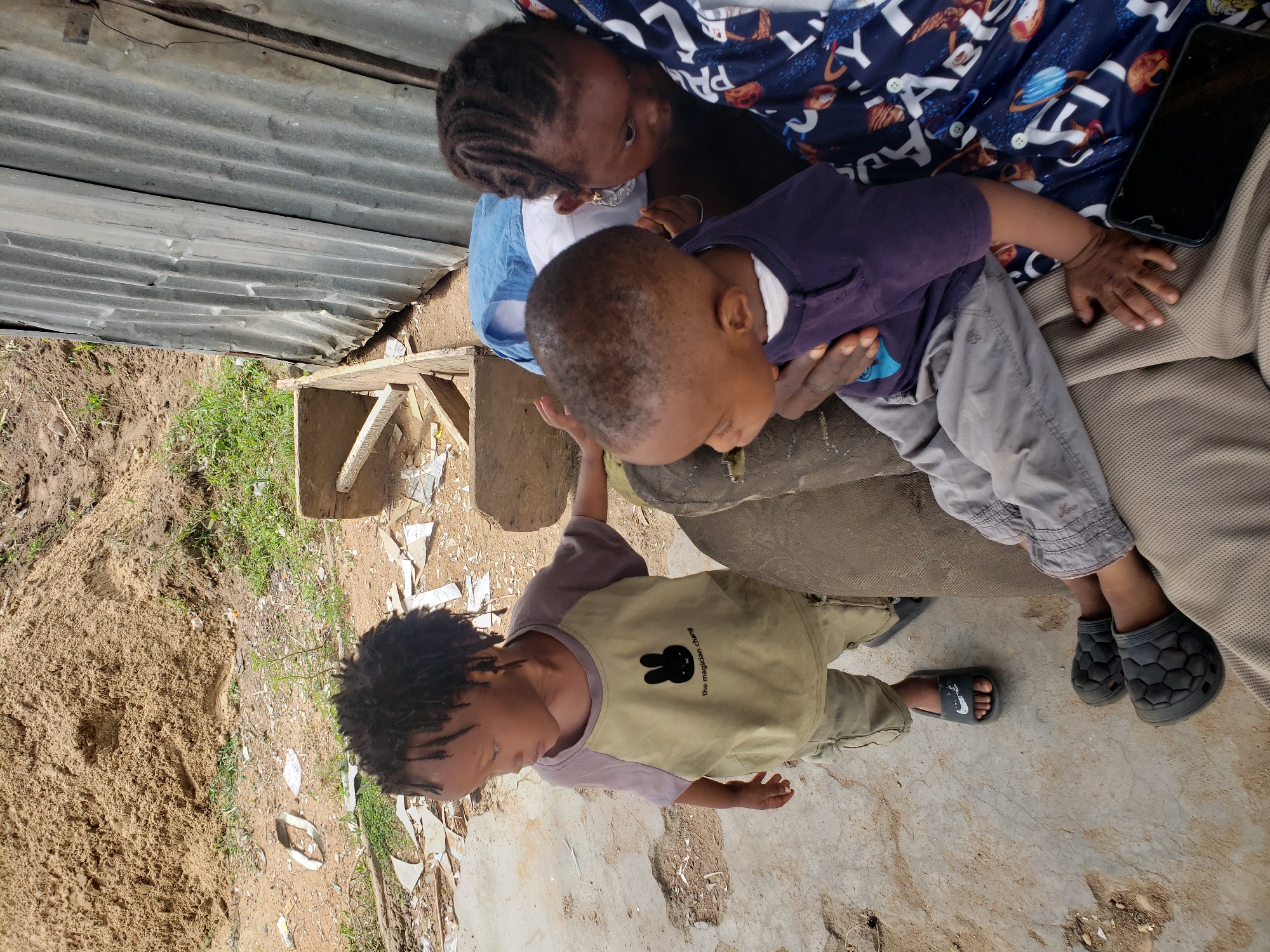 The image, taken in Adankolo, Nigeria, depicts three children in an outdoor, semi-urban or rustic setting during bright daylight.
In the center, an older child, possibly a girl, with dark braided hair and wearing a patterned blue and white shirt, is seated and holding a younger child. The younger child, a baby or toddler, wears a purple top and grey pants and is looking downwards. A black mobile phone with a cracked screen is visible on the lap of the person holding the younger child.
To the left, a slightly older child, appearing to be a boy with dark dreadlocks, stands beside them. This child wears a two-toned t-shirt, light green/beige on the body with a black bunny logo and the text "the magician club," and purple sleeves. He is wearing light brown or beige pants and a dark slipper with a white Nike swoosh logo on his visible foot. The child being held is wearing dark, hexagonal-patterned closed-toe sandals.
The background features a corrugated metal wall or fence on the left side. The ground beneath the children is a mix of dirt, concrete, and scattered debris including broken pieces and plastic. To the far left, a large pile of sandy dirt is visible, along with some green vegetation. A simple wooden structure or bench is also present in the middle ground.