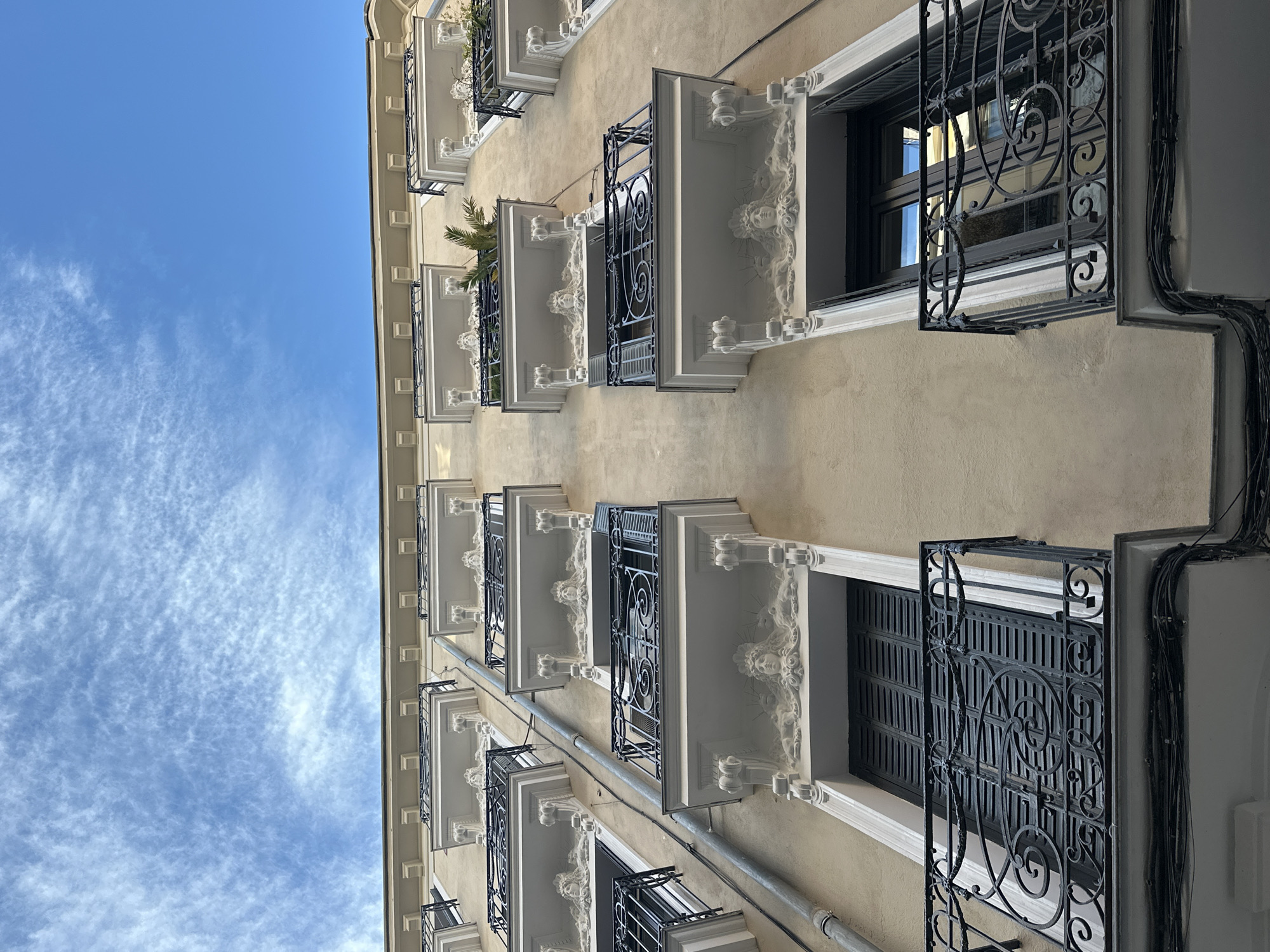 An upward-angled shot of an apartment building in Madrid, Spain, displays a pale yellow facade, rows of white-framed windows, and wrought-iron balconies. Architectural details include ornate carvings on the window frames. Some balconies hold small plants. A drainpipe runs vertically along the building. The sky is blue with scattered clouds.