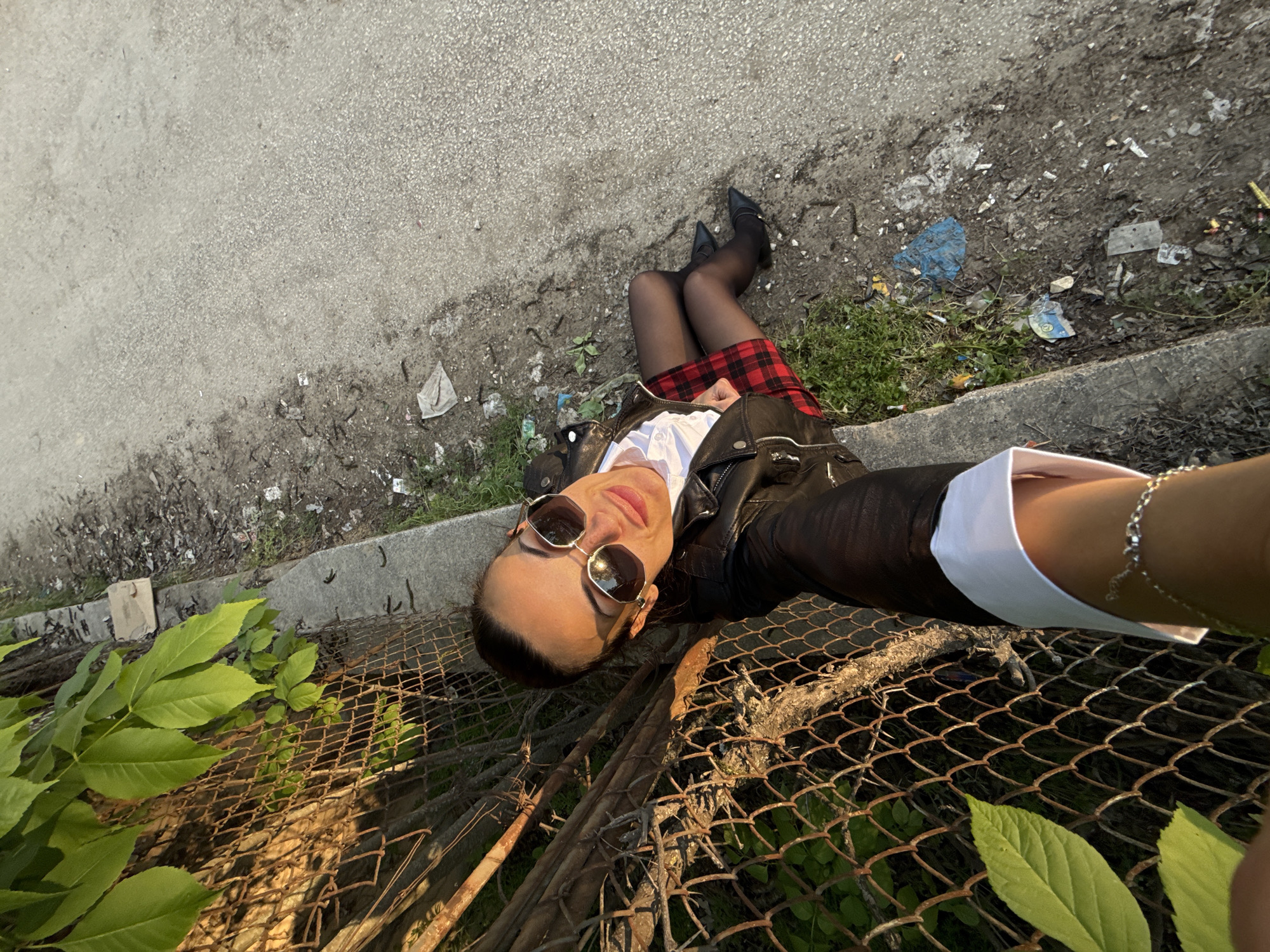 A woman in a black leather jacket, white shirt, and plaid miniskirt poses for a selfie. She wears black tights and heels, and sunglasses. She is positioned near a chain-link fence with greenery, next to a cracked asphalt surface with litter. The photo's location is inferred to be Pleven, Bulgaria.