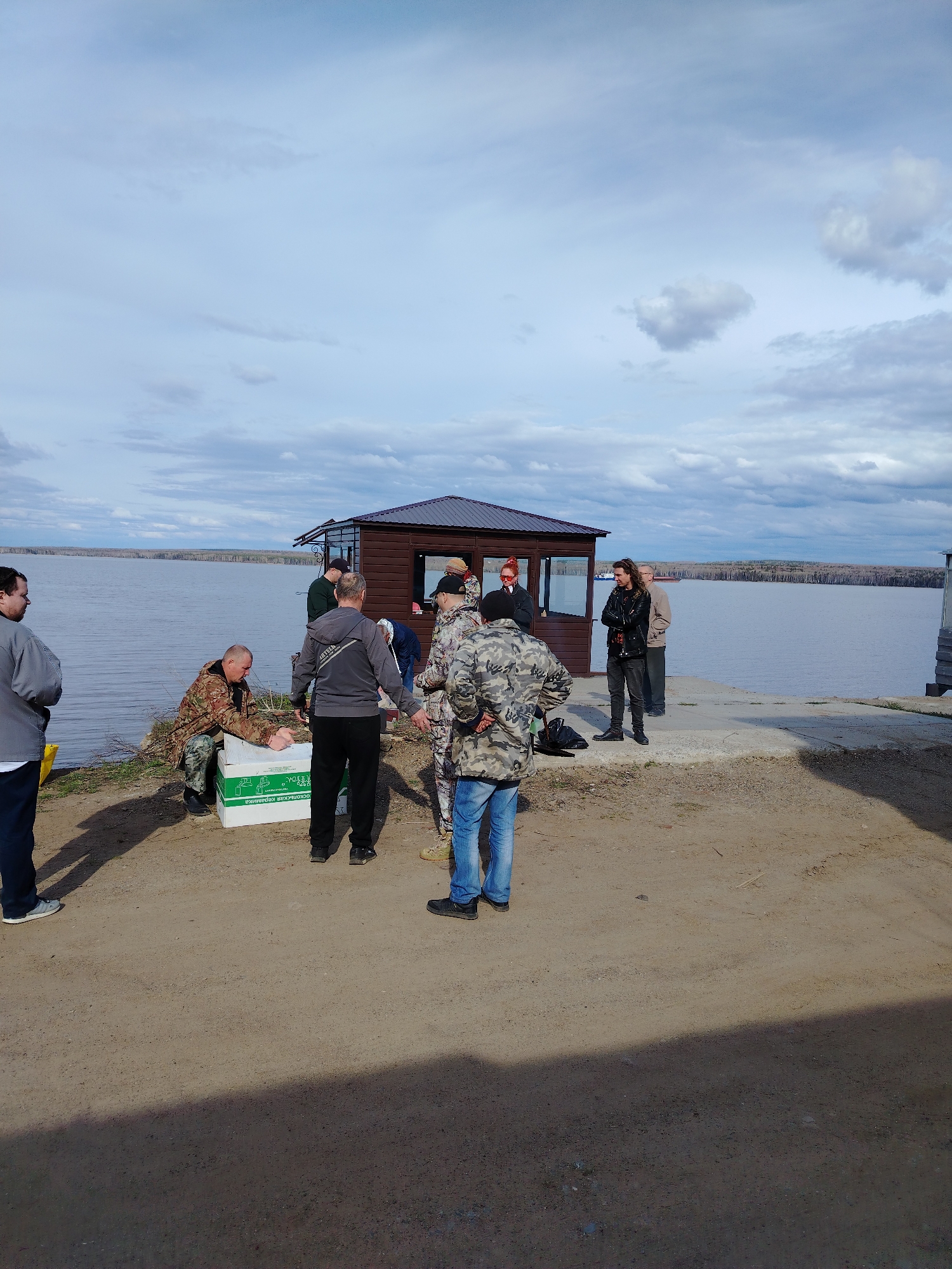 A group of approximately ten people stand on a sandy/dirt area near a body of water. A small, brown, hexagonal-shaped building with a black roof is behind some of the people, adjacent to a concrete pier. Some of the people are clustered around a white box, while others are standing near the building or pier. The background includes the water and trees on the opposite shore, under a cloudy sky.
The box has Russian text on it. Based on the text, the location is likely related to the city of Chaikovsky, Russia.