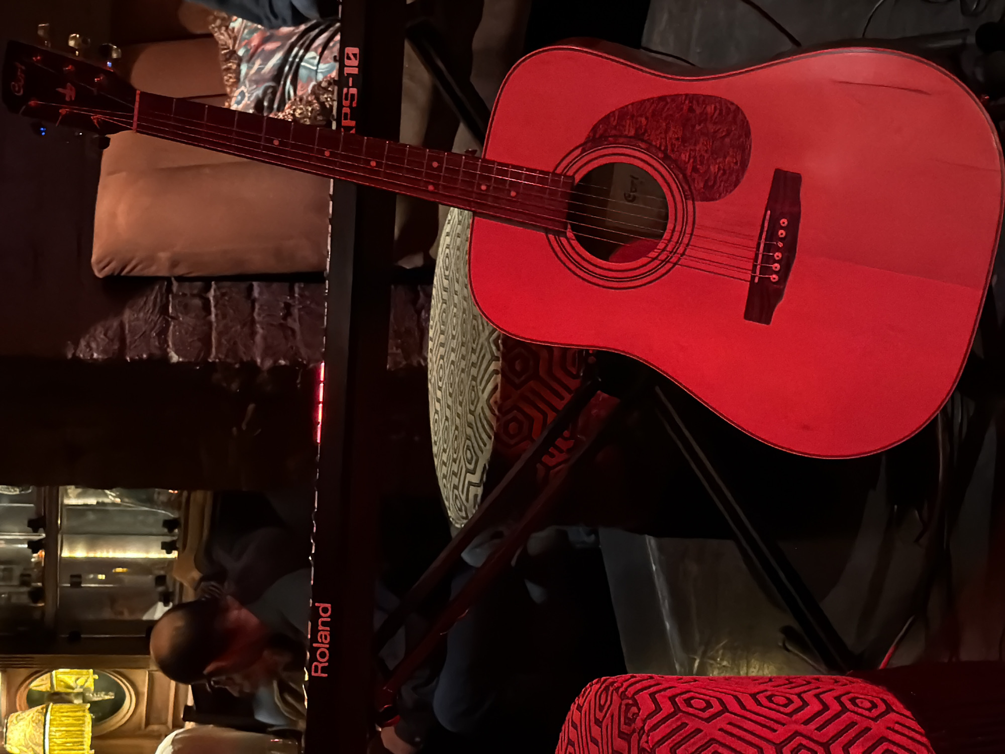 A red-lit acoustic guitar is propped on a stand in a dimly lit location. The guitar is a Cort brand instrument with a dark brown pickguard. The stand is black and positioned in front of a patterned stool. A Roland brand keyboard stand is partially visible to the left of the guitar. In the lower left, a person with a shaved head is bent over, possibly working on some equipment. A shelf with various electronic items is behind them. The overall lighting is subdued and reddish. The city of Cape Town, South Africa, is not discernible from the image.