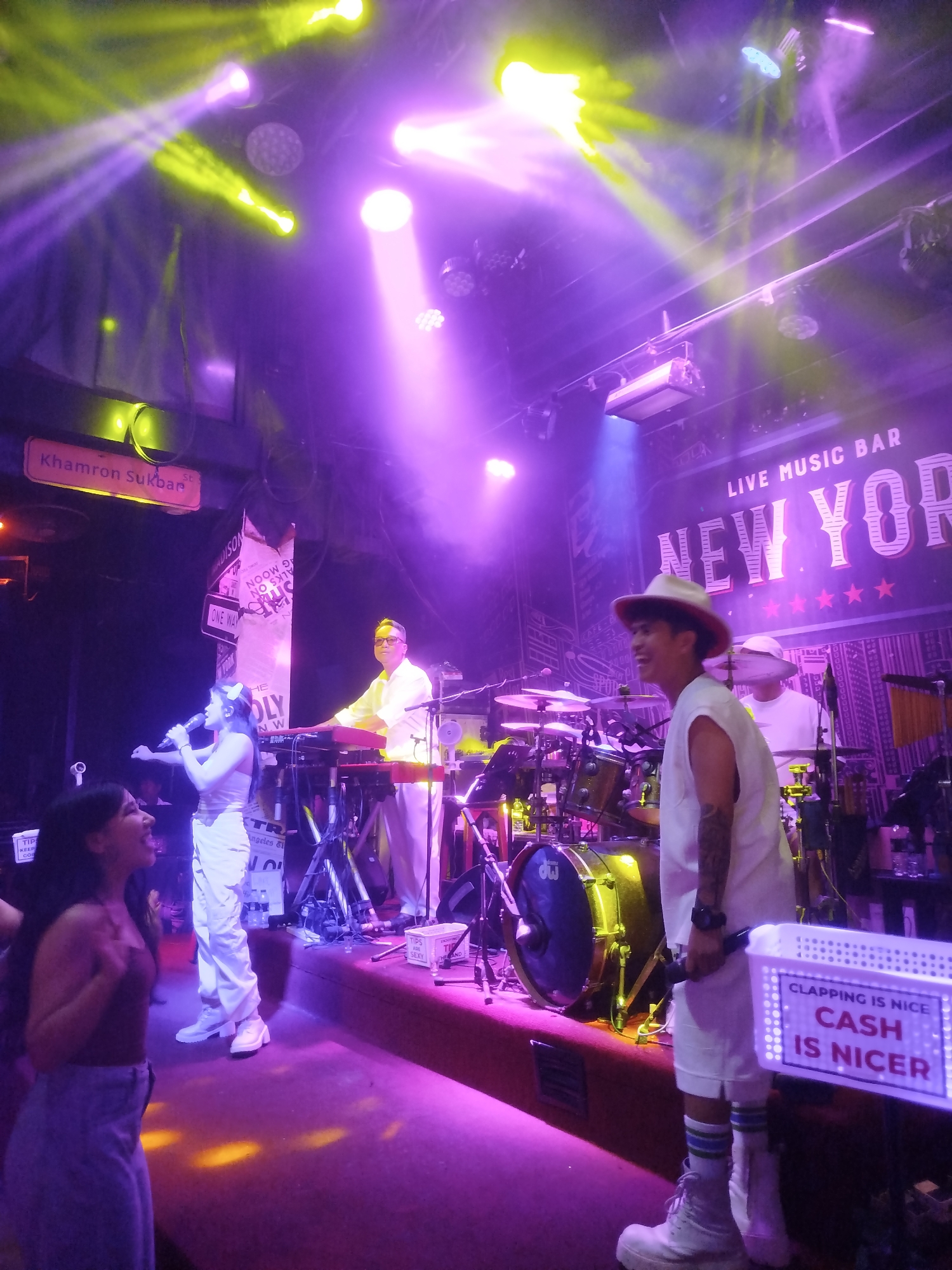 In Patong, Thailand, a live music bar called New York is hosting a performance. The stage is illuminated by purple and yellow lights, creating a vibrant atmosphere. A female singer, dressed in white, holds a microphone and sings to the audience. Beside her, a male keyboardist, also in white, plays a red keyboard. In the background, a drummer is seated behind a drum set. A male performer in a white hat and sleeveless shirt, holding a microphone, stands in the foreground, facing the audience. A sign in front of him reads "CLAPPING IS NICE CASH IS NICER." Two women are visible in the lower left of the image, one singing and the other watching the performance. The scene suggests a lively and energetic evening at a music venue.