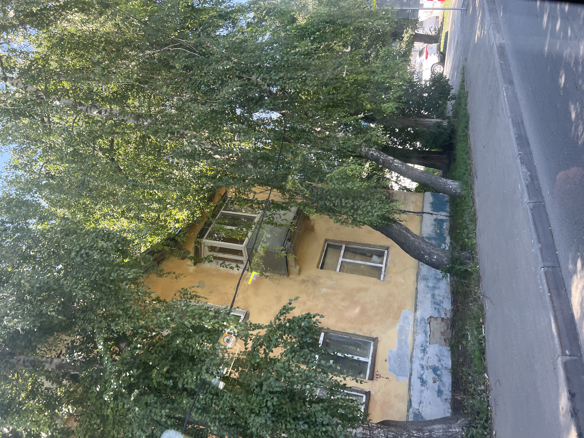 The image shows a weathered, yellow apartment building with a damaged facade, bordered by mature trees with abundant green foliage. The building features several windows, some with white frames and others with clear glass, and a small balcony. The lower part of the building's wall exhibits peeling paint, revealing patches of blue and brickwork. A sidewalk runs along the building, separated from a paved road by a curb. The scene is set on a sunny day, with dappled sunlight filtering through the leaves. A white SUV is visible parked on the side of the road in the background, suggesting an urban or suburban setting. Based on the provided context, this scene is located in the city of Ulyanovsk, Russia.