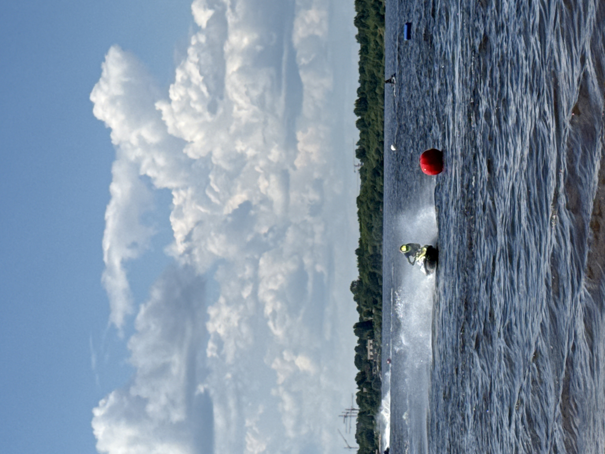 A man rides a yellow and black jet ski on a wavy, dark blue body of water under a cloudy, blue sky. He is creating a white spray behind him as he speeds across the water. In the background, a treeline separates the water from a hazy, urban landscape with tall cranes. To the right of the jet skier, a red buoy floats on the water. The overall scene appears to be a recreational activity in an outdoor setting, possibly during a race or competition.
