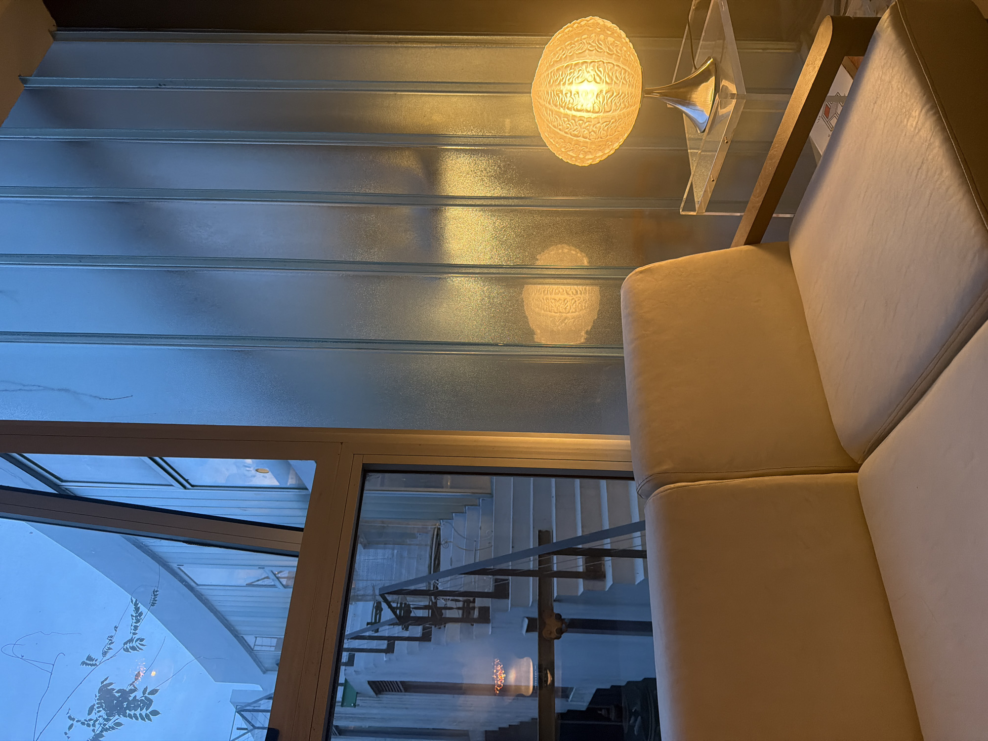 The image captures an interior scene with an unusual perspective, potentially looking down. The focus is on a cream-colored sofa in the right portion of the frame, partially obscuring the view. To the left of the sofa, a window reveals an exterior view of a building, possibly a residence, with greenery visible outside. Adjacent to the window, there's a reflective surface showing an image of a staircase. Above the staircase reflection, a frosted glass-like structure with linear patterns spans across the upper part of the image. A round, illuminated lamp hangs near this frosted structure, adding a warm glow to the scene. The scene does not provide explicit clues that can identify the location to the city of Buenos Aires, Argentina.