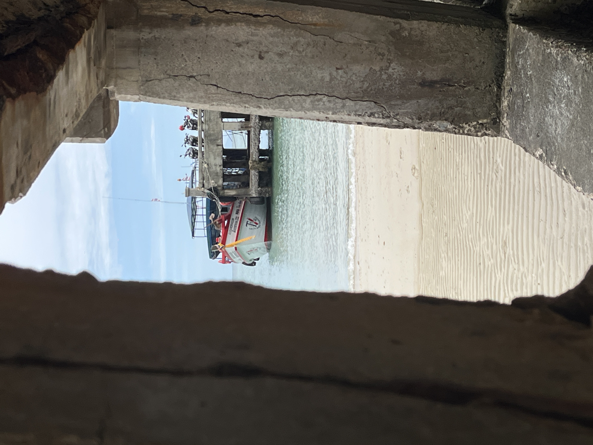 The image shows a view from an opening in a concrete structure, looking out towards a beach and the sea. In the foreground, parts of a rough, textured concrete pillar and wall are visible on both the left and right sides, creating a frame for the scene.
Beyond the concrete structure, a sandy beach with visible raked patterns stretches towards the water. The sea is calm with small waves gently lapping the shore. In the middle ground, a pier or jetty extends into the water. On the pier, several motorcycles are parked, some with riders. A boat, painted white with red and yellow accents and the number "14" visible on its side, is docked next to the pier. The boat appears to be a fishing vessel or a passenger boat.
The sky above is partly cloudy with a light blue hue. The overall scene is one of a coastal area, likely a beach with a pier, in a location that appears to be a popular spot for locals, as indicated by the parked motorcycles. The specific location mentioned, ตำบลเกาะพะงัน, Thailand, suggests this is a tropical island setting.