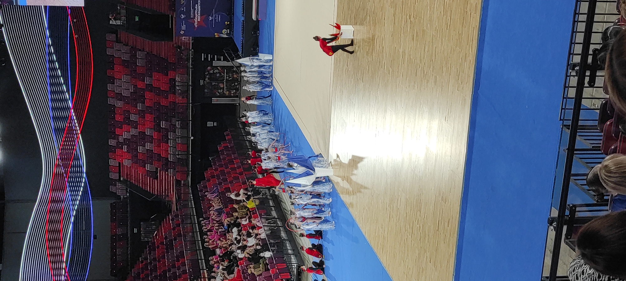 In Moscow, Russia, an artistic gymnastics competition is underway in a large arena. The stands, filled with spectators, are visible in the background, upholstered in red and dark maroon. Above the spectators, a decorative light installation features undulating bands of white, red, and blue lights. On the blue matting of the competition area, a group of young athletes dressed in white and blue costumes stand in formation. To their right, on the wooden floor, a lone performer in a red and black outfit is interacting with a white cube, from which several red feather-like objects are emerging. Another performer in a red outfit is visible in the far left of the frame, observing the scene. The competition area is illuminated by bright lights, creating reflections on the wooden floor.