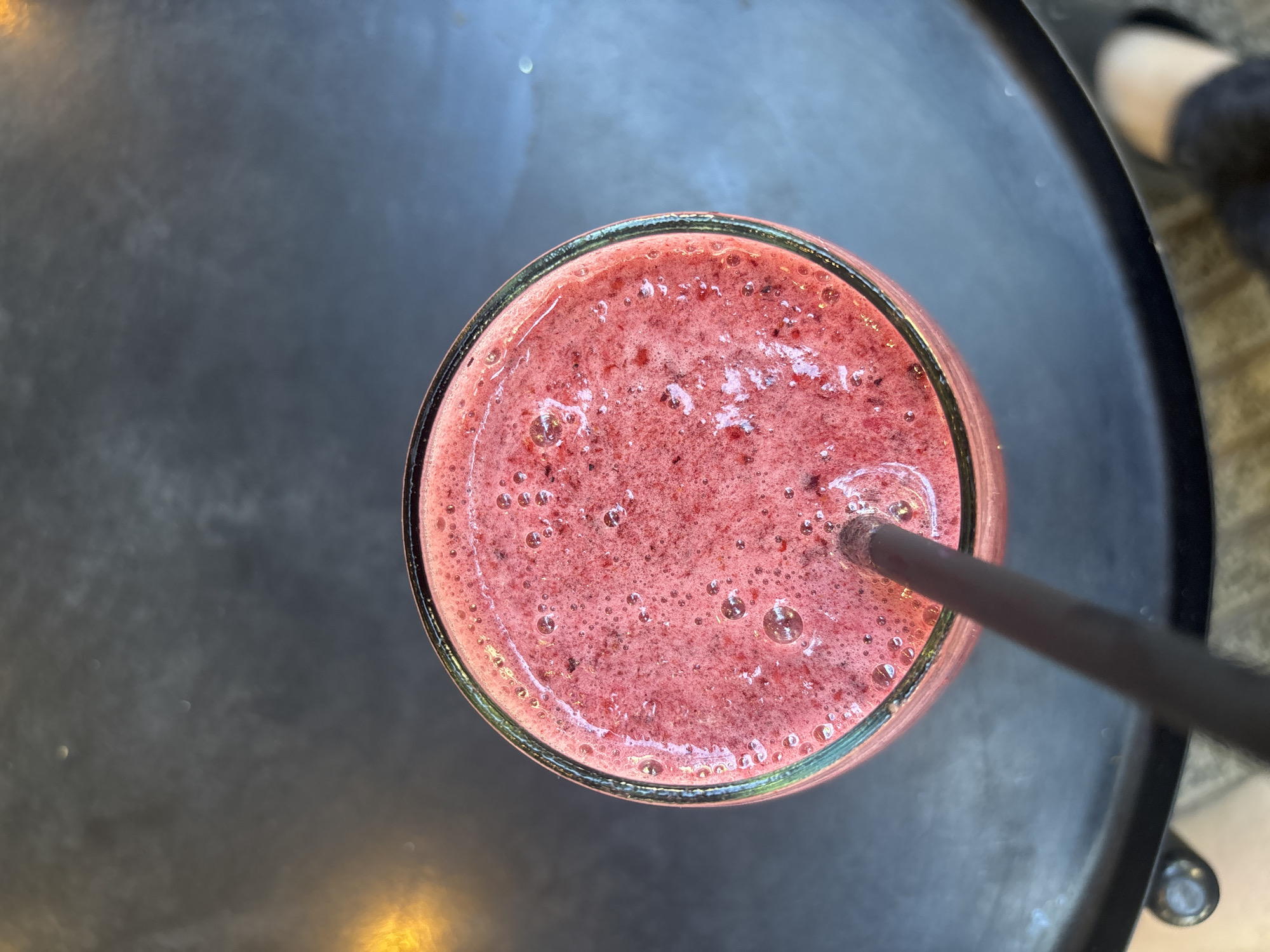 An overhead shot captures a vibrant pink or reddish smoothie in a glass with a dark straw, resting on a dark, circular table. The smoothie, likely berry-based, has a frothy, bubbly texture on its surface, with small dark specks of fruit pulp or seeds visible throughout. The rim of the glass appears to have a subtle green hue. A dark, possibly reusable, straw is inserted into the drink, angled towards the right side of the frame.
The table is a dark gray or black, possibly metal, with subtle reflections indicating light sources from above or around. The surface shows some slight texture or variations in color. In the upper right background, out of focus, is the partial view of a foot wearing a light-colored shoe or sock and dark pants, suggesting the presence of a person seated nearby. Another light reflection is visible in the upper left background on the table surface. The lighting suggests it is daytime. The scene evokes a casual setting, possibly a cafe or outdoor dining area in Buenos Aires, Argentina. There is no visible text in the image.