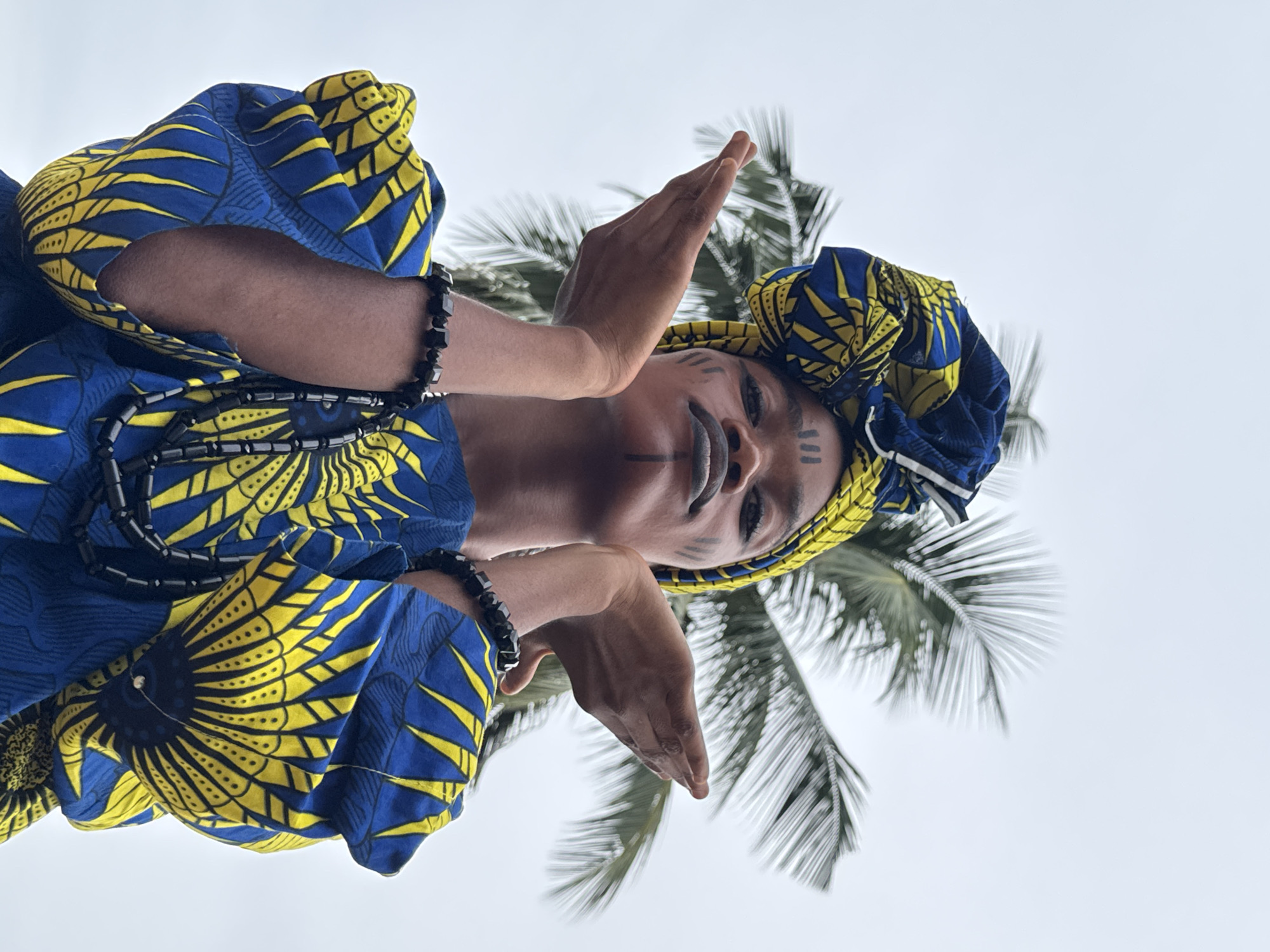 The image shows a person with dark skin wearing a blue and yellow patterned fabric, possibly traditional clothing, along with a matching headwrap. They have dark markings painted on their face. They are wearing black beaded necklaces and bracelets. The person is holding their hands up near their face, with their fingers pointing upwards.
Behind the person is a palm tree with green fronds, and the sky is visible in the background. The camera angle is a low-angle shot, looking upwards at the subject.
While the image may depict a scene reminiscent of Badagry, Nigeria, precise verification of its location within Badagry is not possible without explicit confirmation from the source.