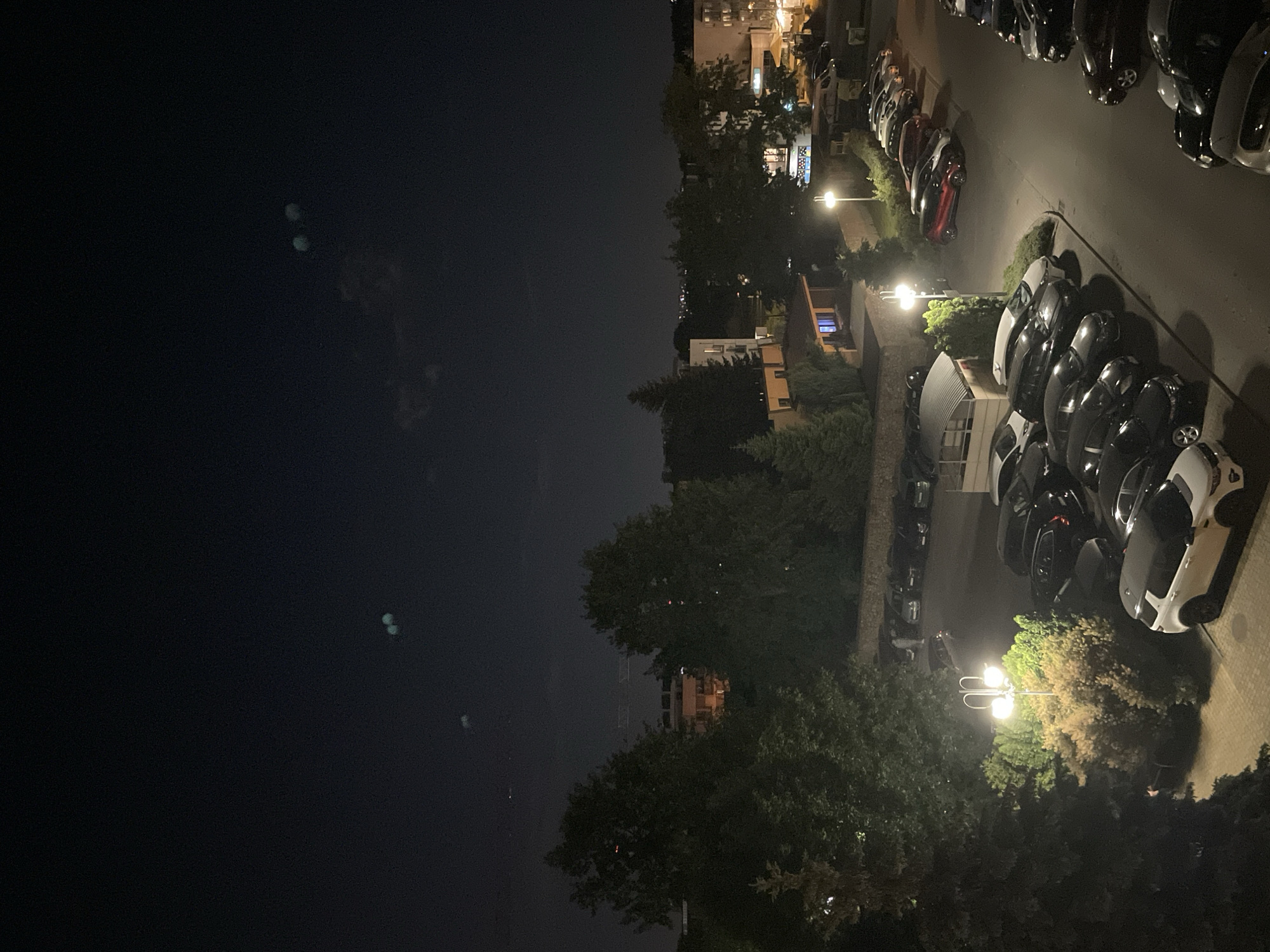The image shows an aerial view of a parking lot in an unknown city at night. The parking lot is lined with parked cars, illuminated by street lights. Trees and buildings are visible in the background. The sky is dark.