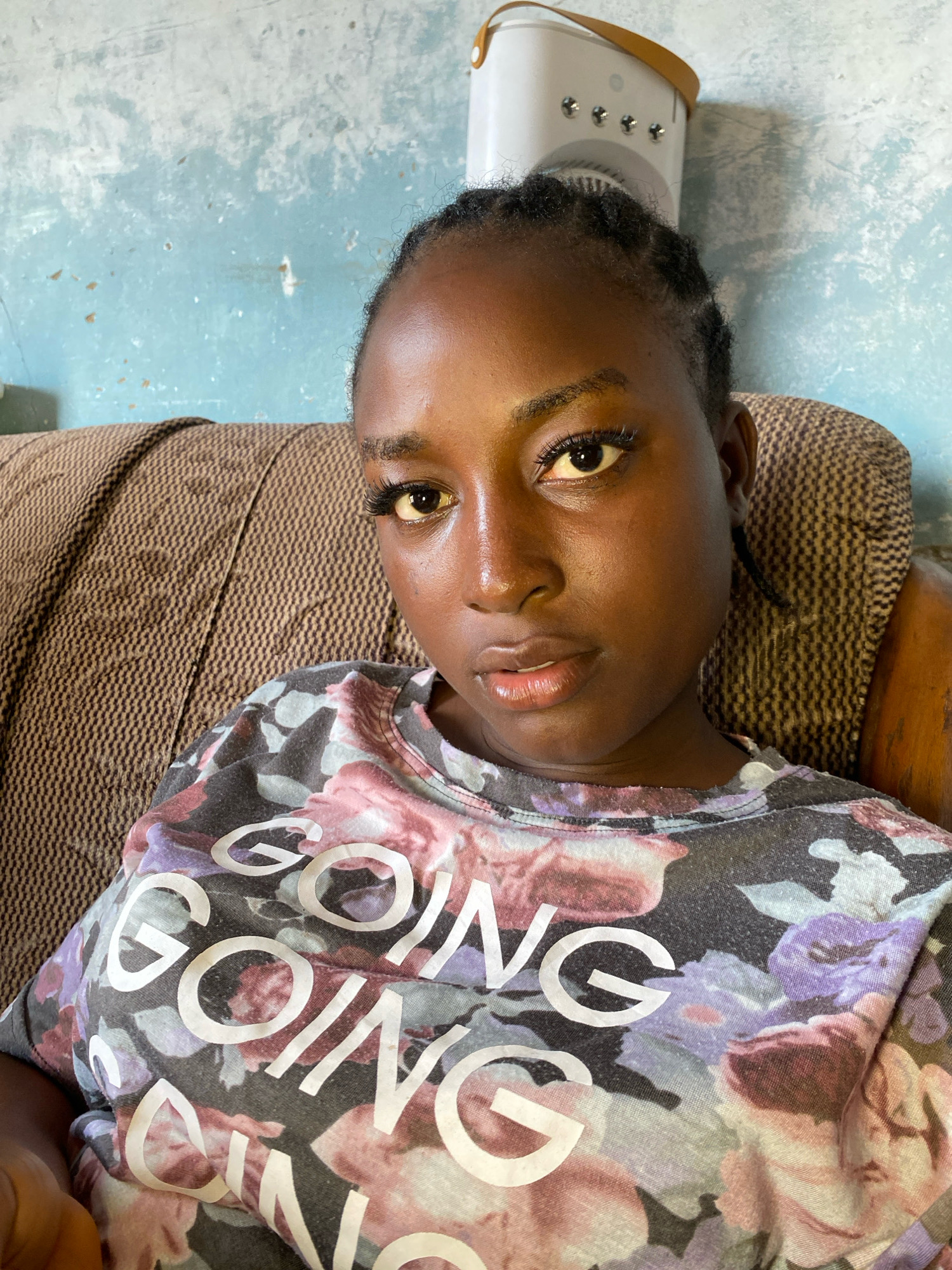 A young Black female is sitting on a patterned brown sofa against a light blue, textured wall. She is looking directly at the camera, her expression neutral. Her hair is styled in thin braids, and she is wearing a t-shirt with a floral pattern and large white text that reads "GOING GOING". A white electronic device with a brown handle is visible behind her, near the top right of the frame. The scene appears to be an indoor setting, possibly a living room, in Sabon Tasha, Nigeria.