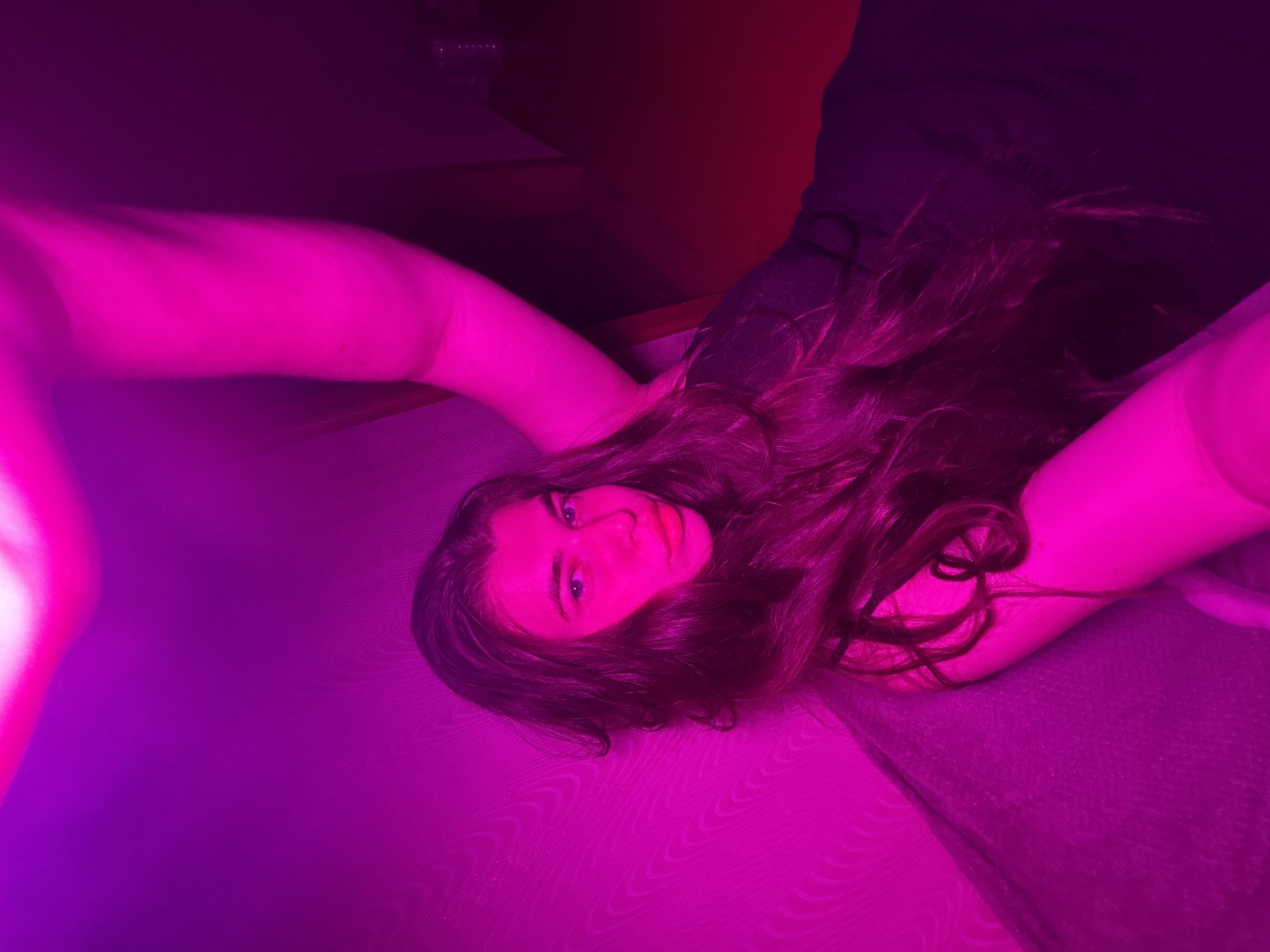 A young woman with long, dark hair is lying down, illuminated by pink and purple lighting. She is wearing a dark garment and her face is turned towards the camera, with her eyes open. The background is blurred, but appears to be a domestic setting. It is not possible to determine the location within the city of Мещёры, Russia from the image alone.
