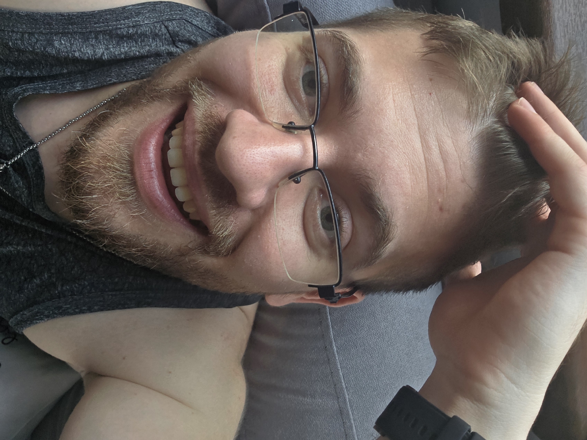 The image presents a close-up, angled portrait of a young adult male lying down indoors, likely in Yagodovo, Bulgaria. He is positioned with his head tilted towards the left of the frame, looking directly at the camera with a wide, open-mouthed smile, revealing white teeth. The man has light brown hair, a light beard or stubble, and green or hazel eyes. He is wearing thin, black-framed glasses.
He is dressed in a dark grey, heathered sleeveless top or t-shirt, and a thin, metallic ball-chain necklace is visible around his neck. His left hand is raised, with fingers lightly touching his hair on the side of his head. A black digital wristwatch is worn on his left wrist. The setting appears to be a comfortable indoor space, indicated by the grey upholstered surface, possibly a couch or bed, on which he is resting. The lighting is bright and diffused, suggesting it is daytime. The man's expression conveys happiness and relaxation. Small red spots are visible on his forehead. No visible text is present in the image.