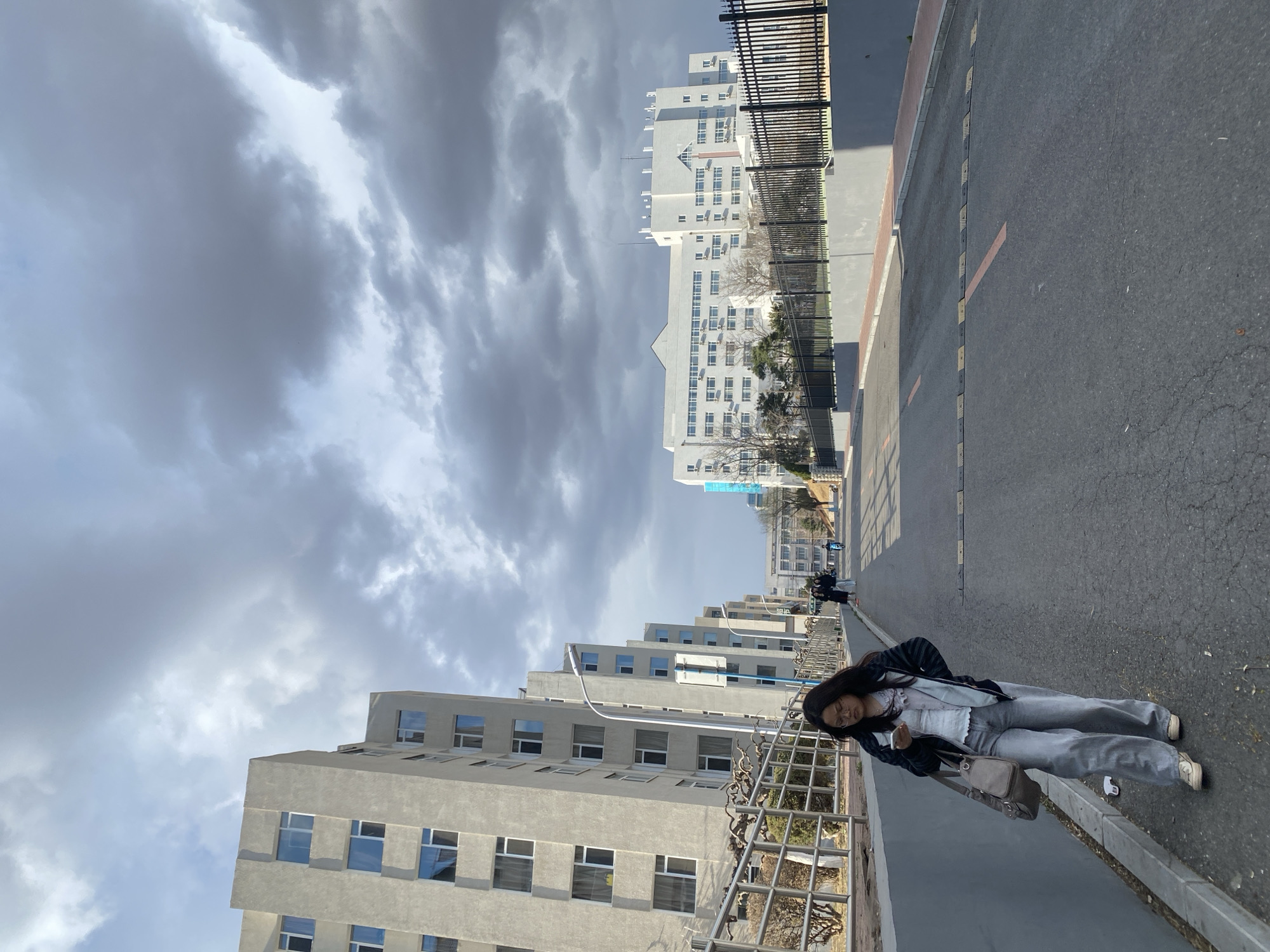 A full shot of a woman standing on an asphalt road in Zhongtun, China. Several white apartment buildings line the left side of the road and are parallel to a tall white building with multiple small windows on the right side. The sky above is overcast with grey clouds. The woman is wearing grey pants, a white top, and a black jacket, and is holding a grey purse. A grey metal fence runs along the left side of the road. Other people are seen walking further down the road in the distance.