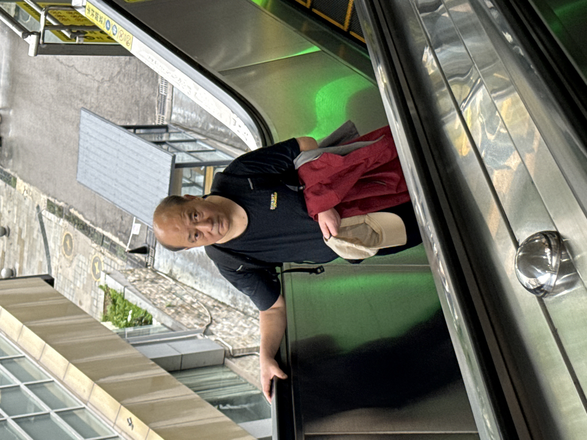 A man is riding an escalator. He is holding a hat and a jacket. The escalator is green. The location appears to be the Nan'an District of China, based on the proximity of the escalator to buildings and street-level scenery.