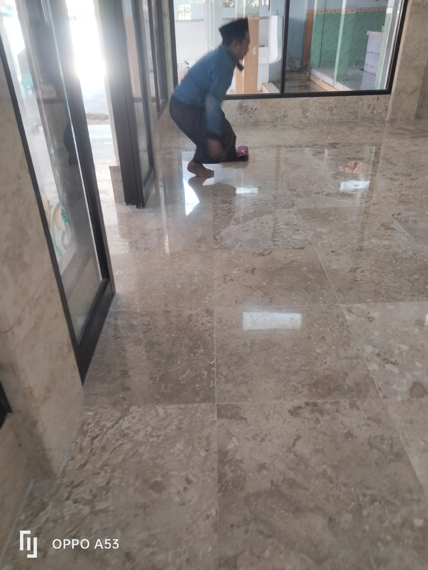 A person is shown in a mosque in Badung, Indonesia. The person is wearing a blue shirt and black pants and is in a kneeling position. The floor of the mosque is made of marble tiles and is shiny. The person is looking down at the floor. The background of the image shows glass doors and windows, with a reflection of the outside world visible. The overall scene suggests a moment of prayer or contemplation within a religious setting.