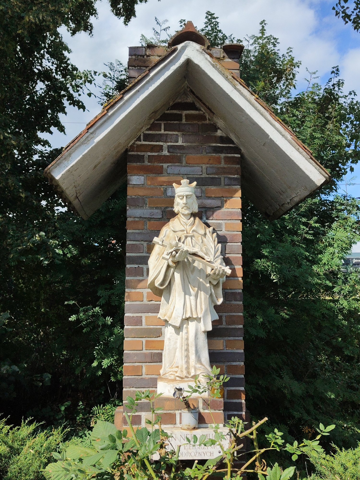 A religious shrine with a statue of a male figure is positioned outdoors, partially sheltered by a peaked roof structure. The shrine is constructed of brick and houses a white stone statue depicting a robed man with a beard and a crown, holding a cross and a sprig of leaves. Below the statue, a stone plaque with inscriptions is visible. The shrine is surrounded by dense green foliage and trees, with a partially visible blue sky and clouds in the background. This shrine is located in Gdynia, Poland.