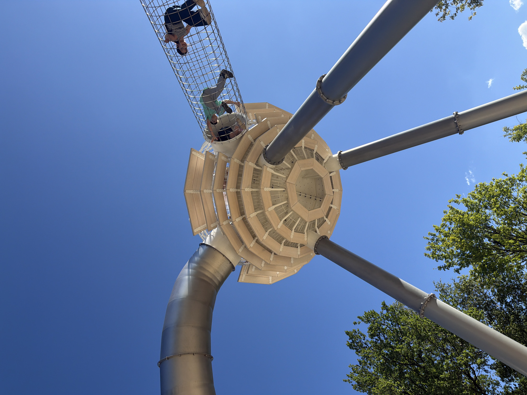 A low-angle outdoor shot captures a modern playground structure against a bright blue sky. The structure is a multi-level, octagonal building supported by three large, gray metal poles. A wide, silver metal tube extends from the left side of the frame, connecting to the top level of the octagonal building. A netted tunnel, also silver, stretches from the top of the building towards the right, with two people visible inside. One person, wearing a striped shirt and dark pants, is at the far right of the tunnel, looking down. Another person, wearing a green t-shirt and gray pants, is in the middle of the tunnel, bending over. The building's exterior is composed of stacked wooden panels forming an octagonal shape. Green trees with visible foliage frame the bottom of the image on the left and right. The scene appears to be a clear, sunny day.