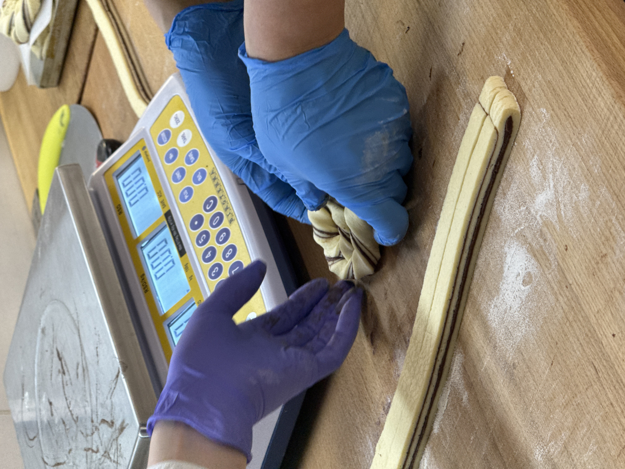 The image shows a person wearing blue gloves manipulating dough with chocolate filling on a wooden surface next to a digital scale. The scale shows "0.00" on its display. A second gloved hand, this one in purple, is reaching toward the dough. The scene appears to be in a food preparation area, possibly a bakery. The location in the city of Vracar Urban Municipality, Serbia cannot be determined from the given information.