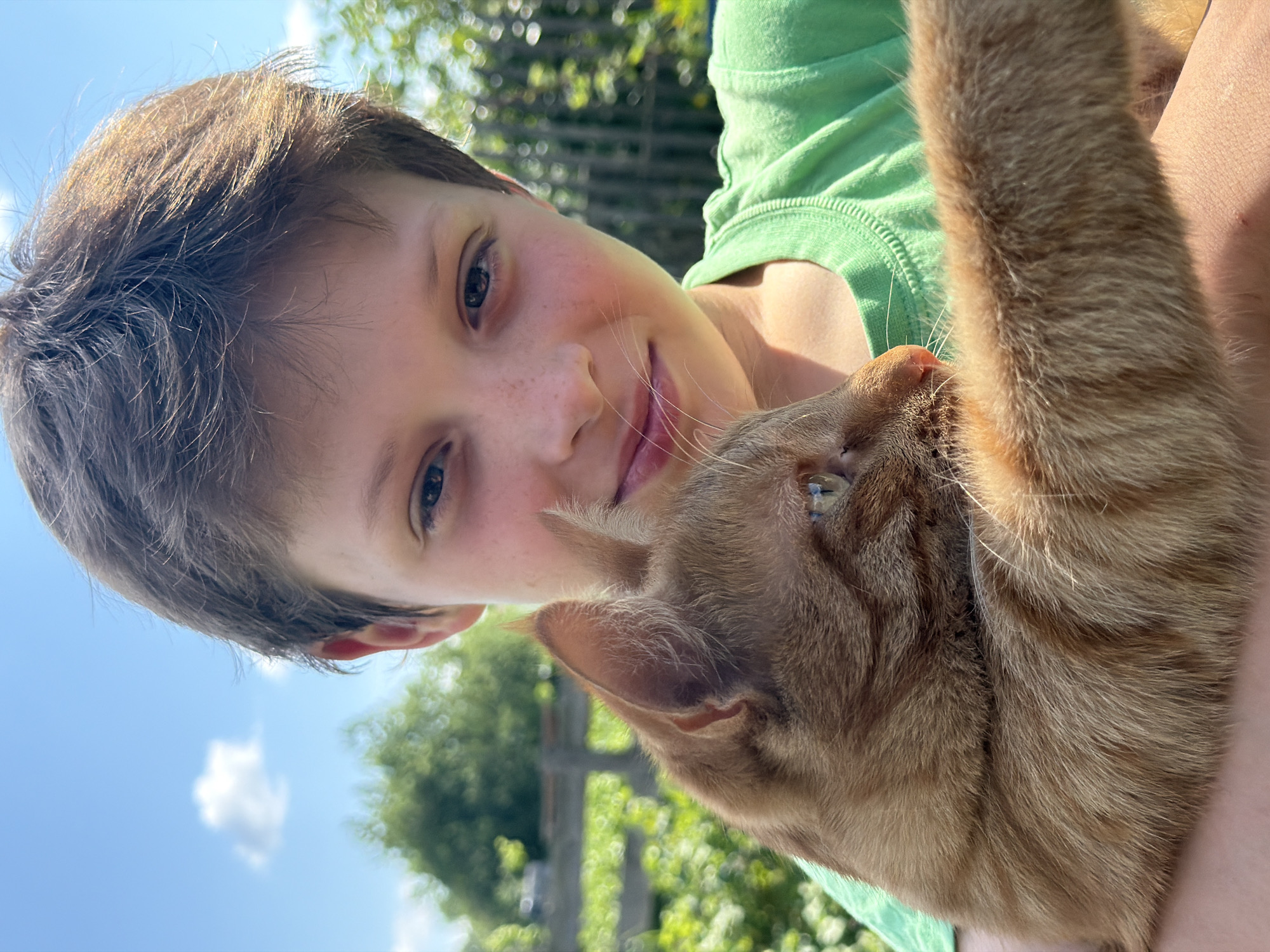 A young boy with light brown hair and freckles is holding an orange tabby cat. The boy is smiling and looking at the camera, while the cat is looking to the side. They are both in an outdoor setting, with a blue sky and some greenery in the background. The image appears to be a selfie, taken on a sunny day. There is no indication of the location in the city of Уральское, Russia.
