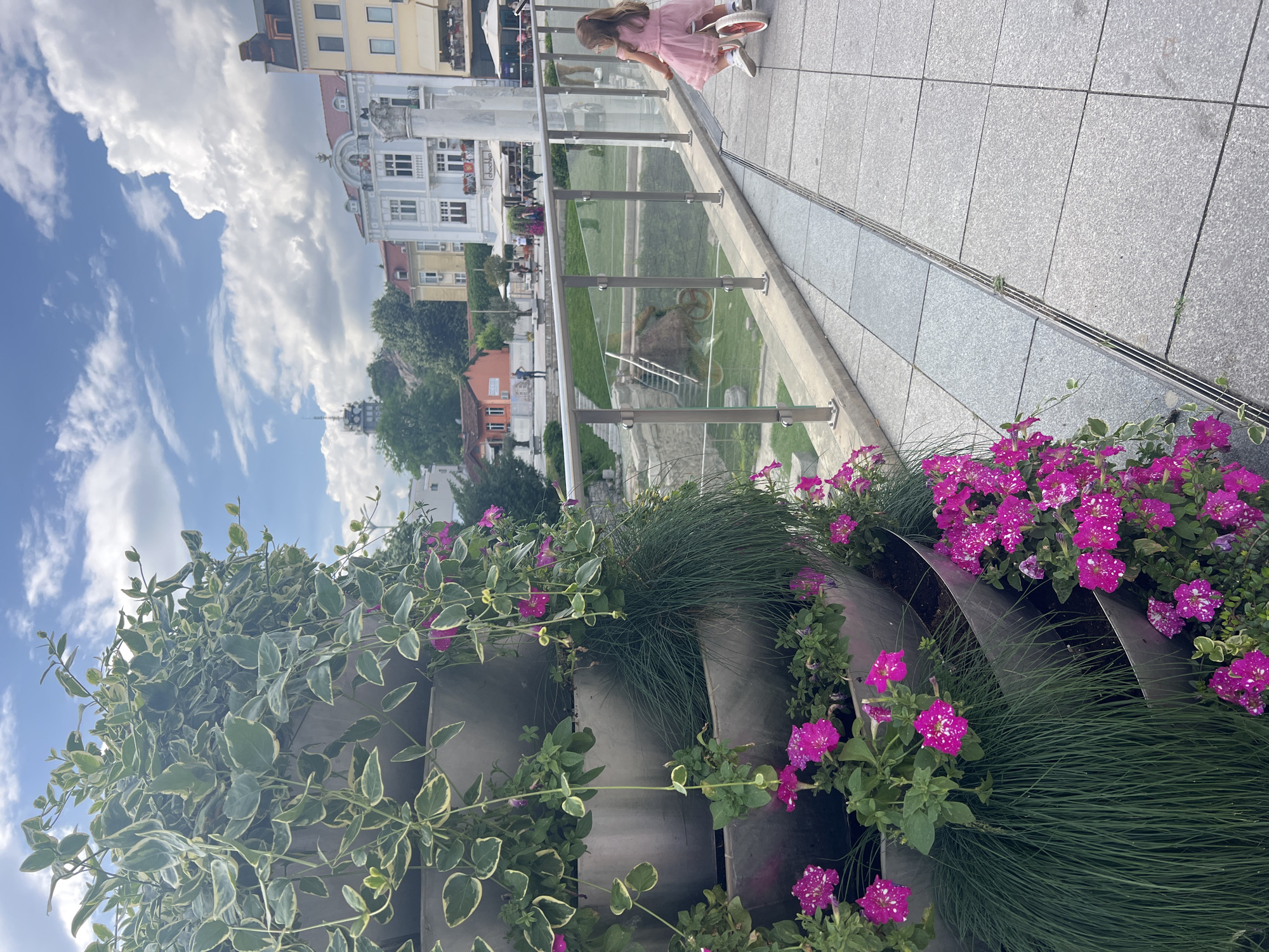 A view from Plovdiv, Bulgaria, shows a young girl in a pink dress kneeling on a tiled sidewalk, peering over a glass railing. Below her is a park area with a decorative sculpture resembling an old-fashioned bicycle with oversized wheels. In the background, there are buildings typical of the urban architecture in Plovdiv, set against a backdrop of a bright blue sky dotted with puffy white clouds. The foreground features tiered planters filled with greenery and vibrant pink flowers with white specks.