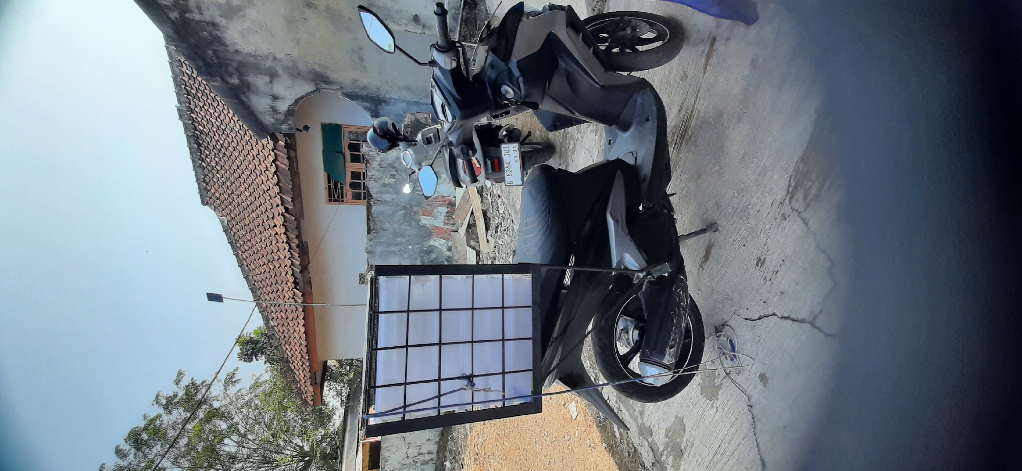 A matte black scooter with a white, grid-like box attached to its rear is parked on a cracked concrete surface. The scooter is positioned in front of a weathered, light-colored building with a red tiled roof. Another scooter, light gray, is parked behind the first. The scooter's license plate reads "B 6264 JU1". The box is partially covered with a white, translucent material and secured with blue ropes. The scene appears to be outdoors, under a clear blue sky, with some greenery visible in the background. The location is identified as Cisauk, Indonesia.